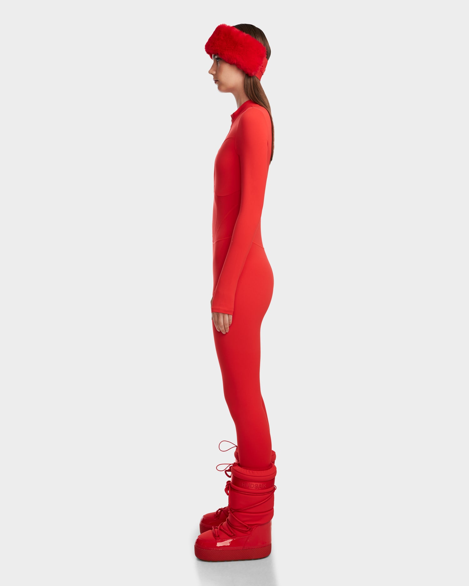 A woman in profile wears the Rudsak IVY - 8524599 Flame Red form-fitting stretch bodysuit, a red faux fur headband, and tall red boots, channeling ski-chic style against a light gray background.