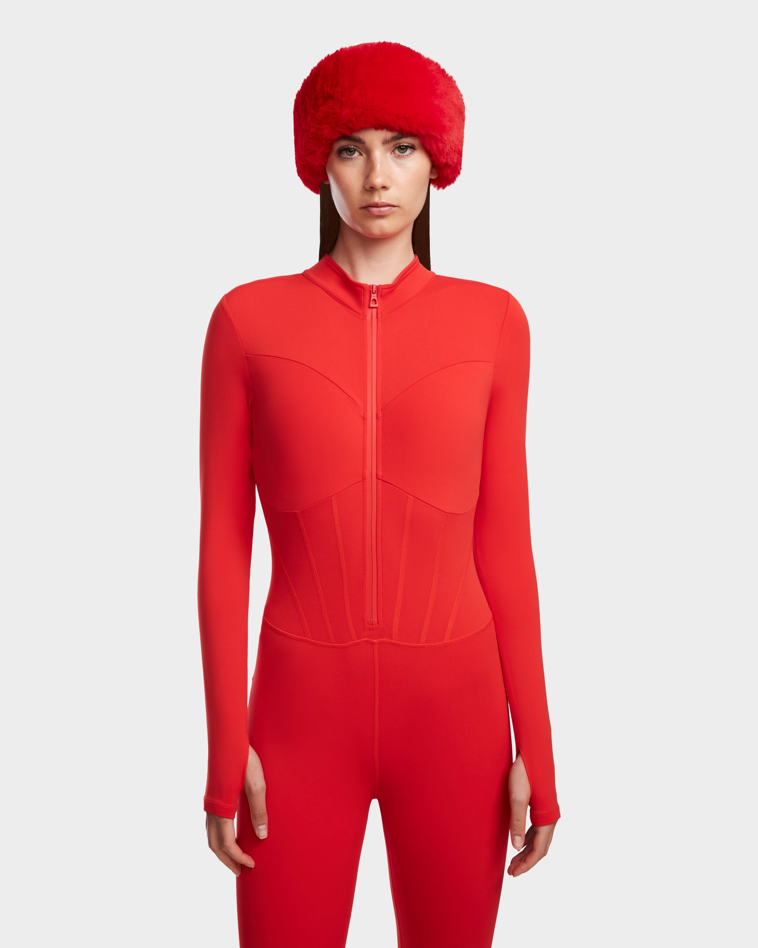 A woman models the Rudsak IVY - 8524599 Flame Red ski-chic zip-up bodysuit with a front zipper and large red hat, set against a plain light gray background.