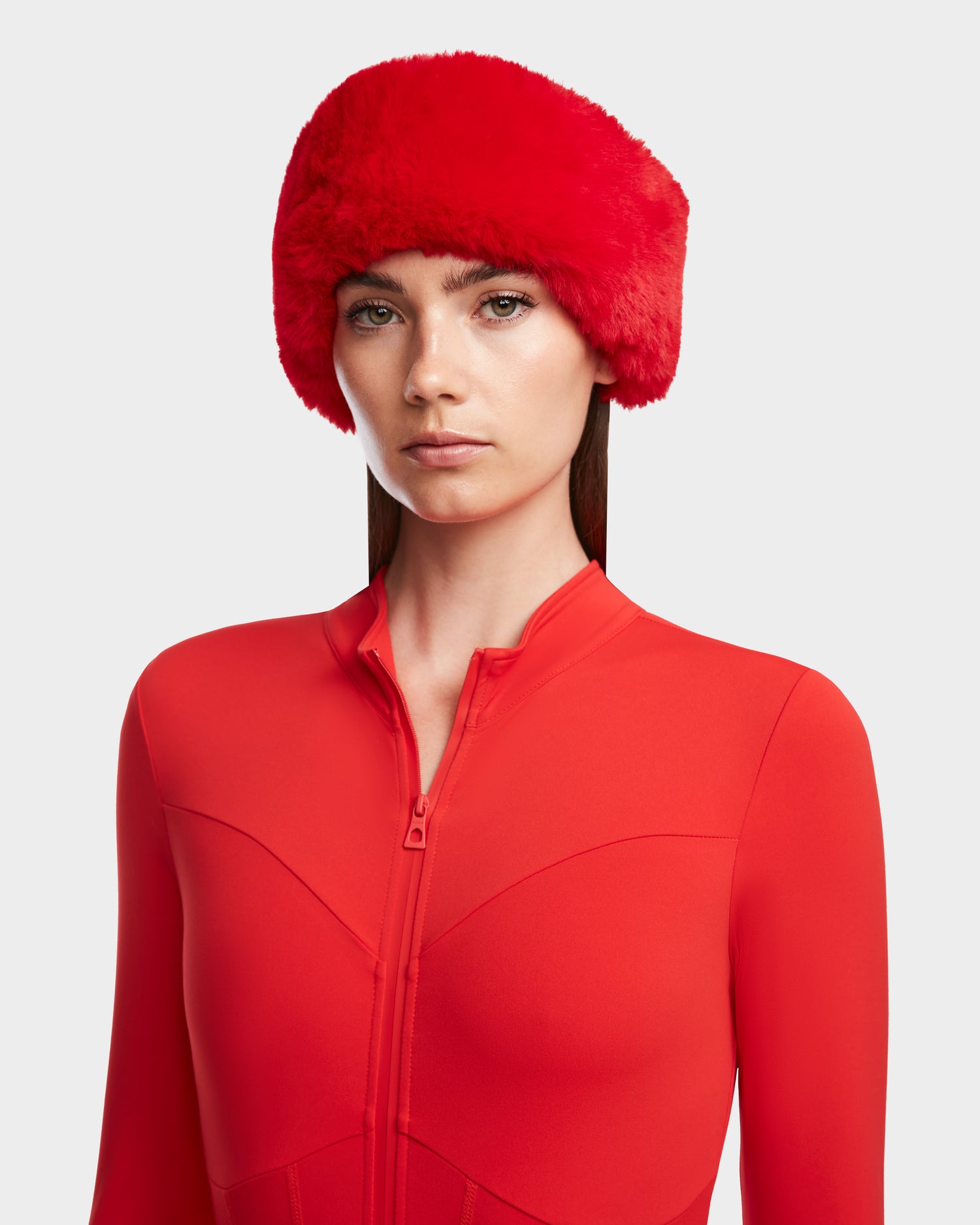 A woman wears the Rudsak IVY - 8524599 Flame Red fitted zip-up bodysuit and a matching faux fur hat, looking directly at the camera with a neutral expression against a plain light background.
