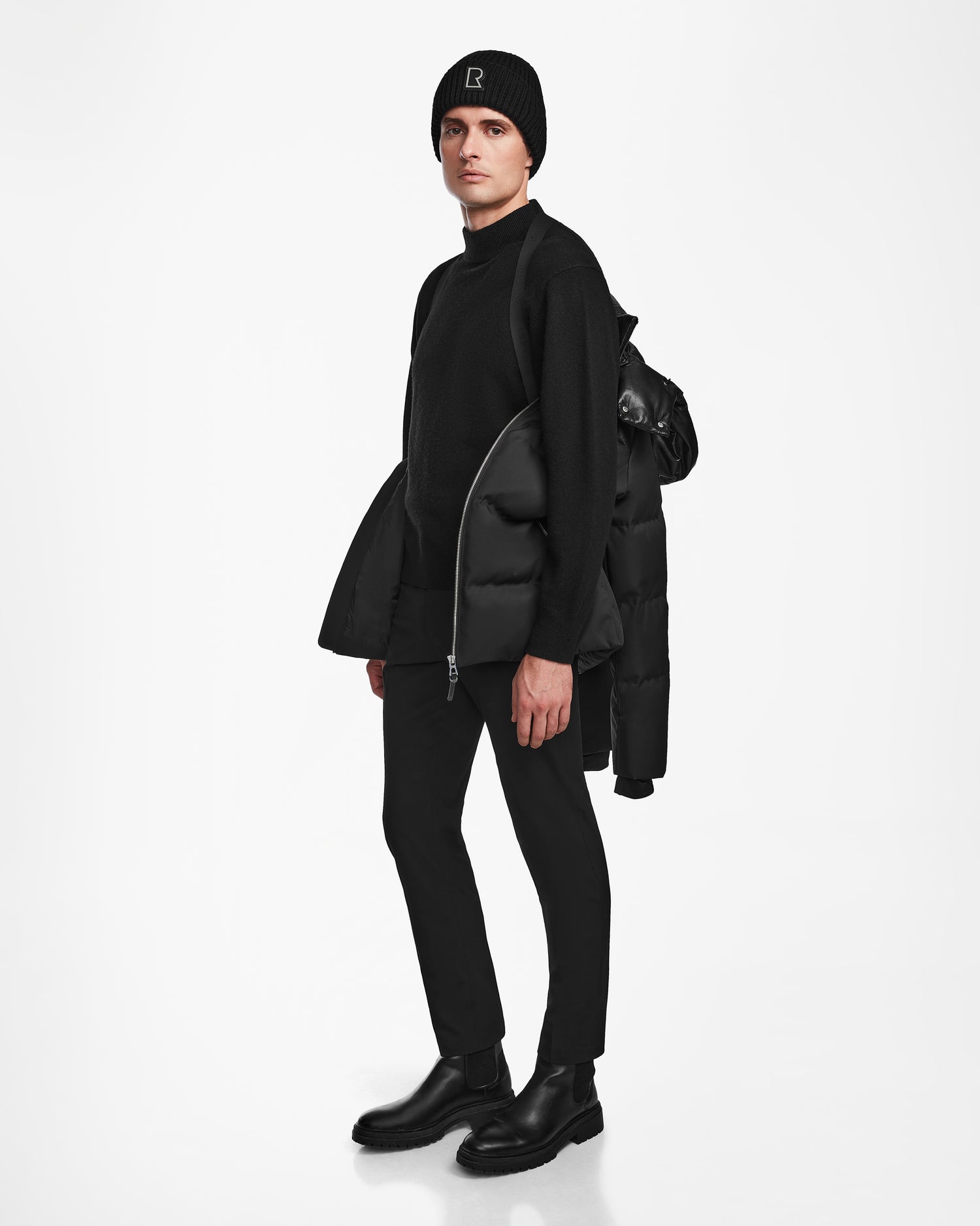 A person stands against a white background wearing a black beanie, sweater, pants, and boots, with the Rudsak JACK - M124507 Black men’s down puffer jacket draped over one shoulder.