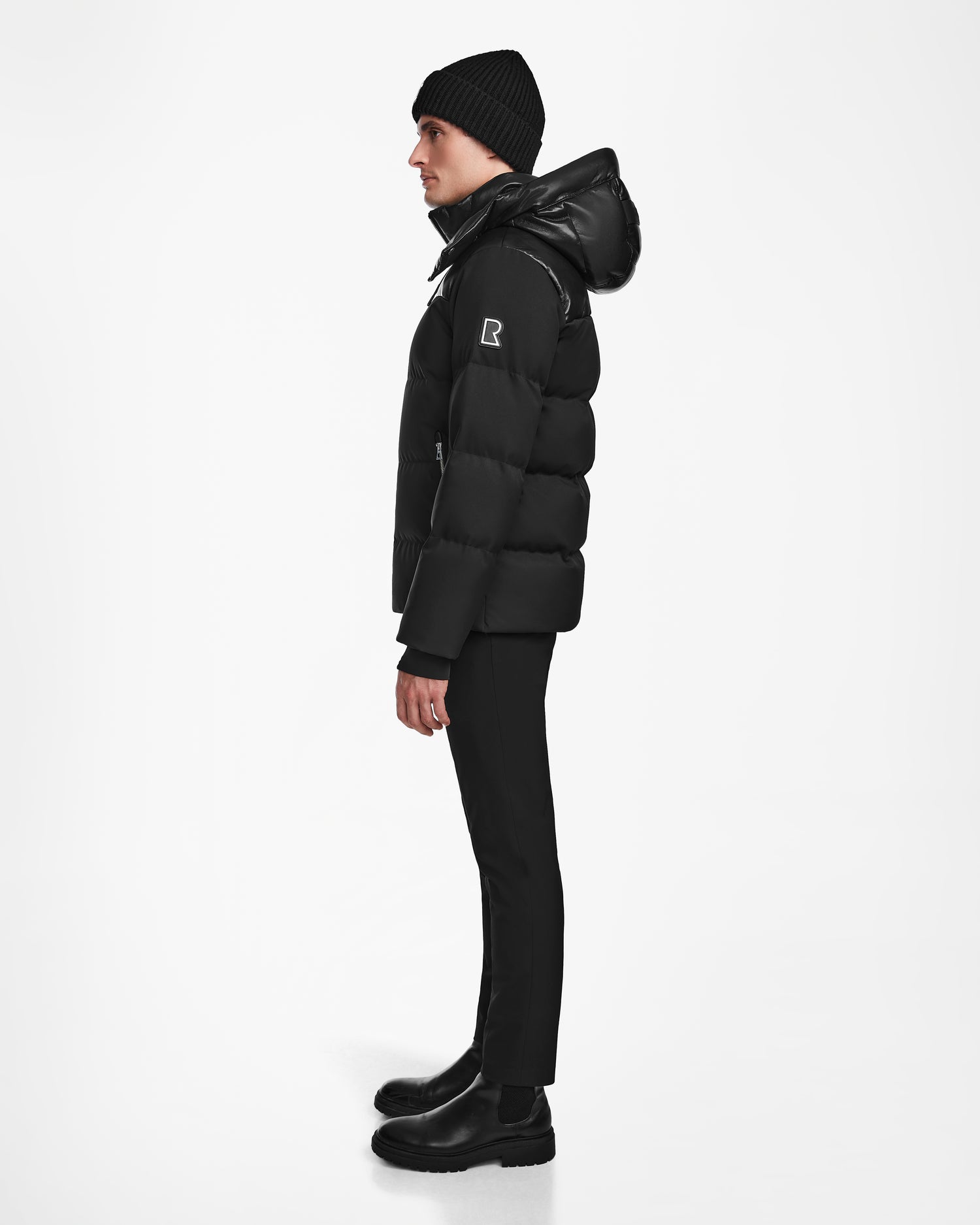 A person in profile against a white background wears the Rudsak JACK - M124507 Black winter jacket, paired with a black beanie, pants, and boots.