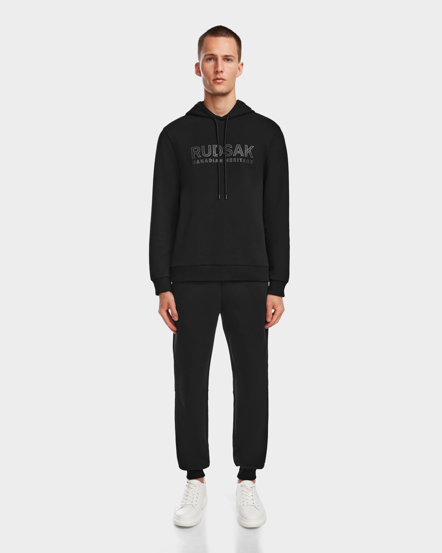 A man faces forward against a white background, wearing the black Rudsak JARED - 6524533 hoodie and matching jogger pants, paired with white sneakers.