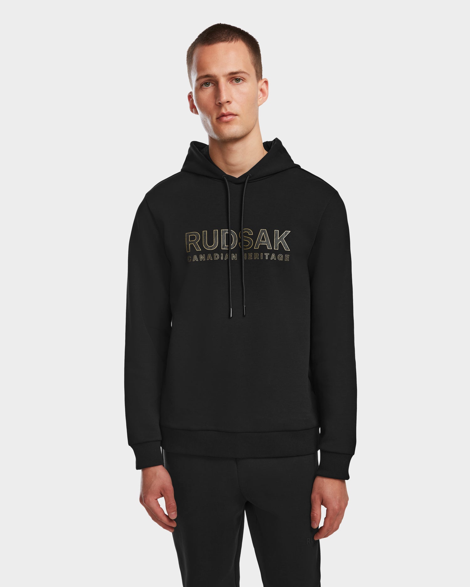 A man stands against a white background, wearing the Rudsak JARED - 6524533 Black mens hoodie, featuring RUDSAK CANADIAN HERITAGE printed in gold on the front.