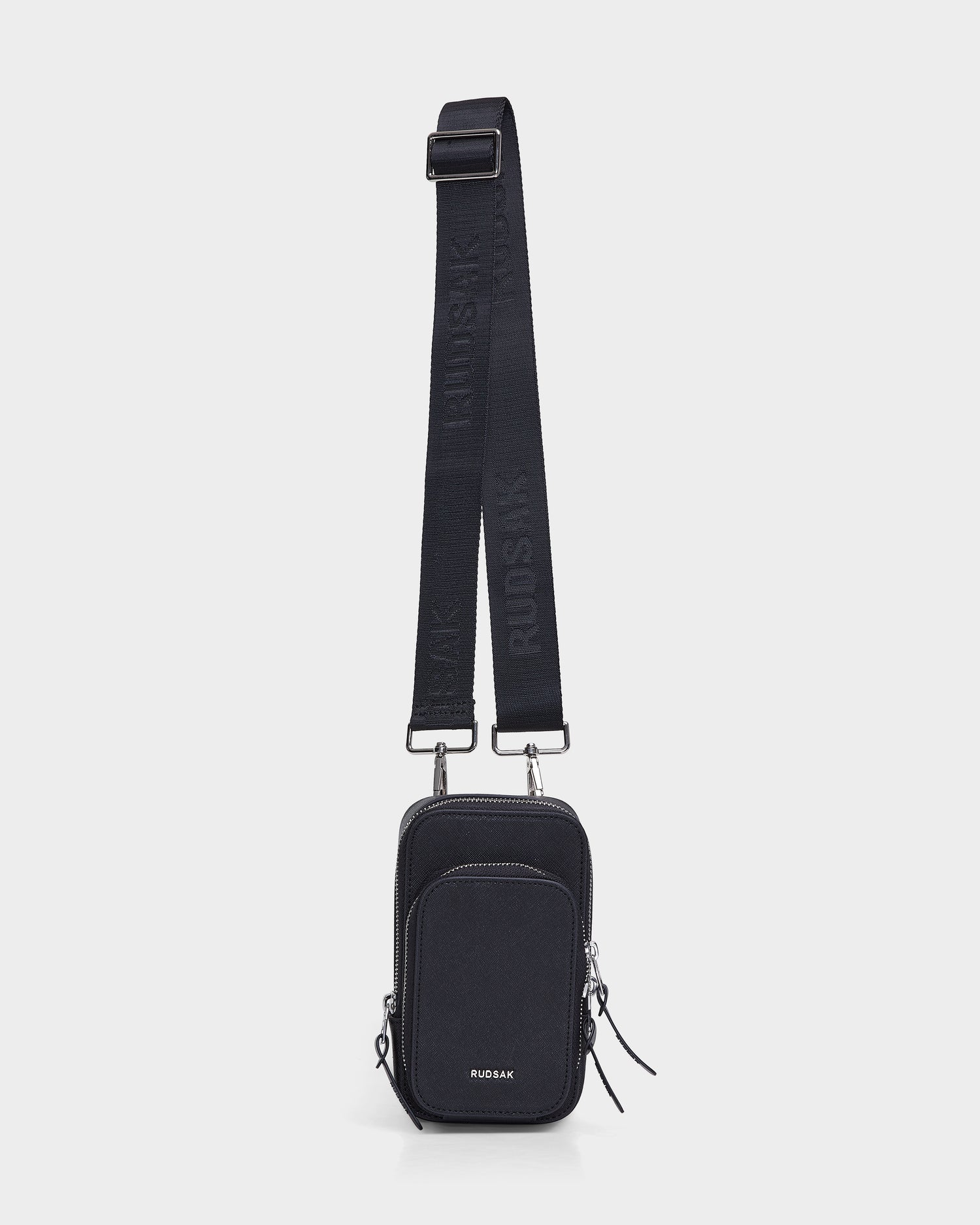 The JIMIE - L324505 Black by Rudsak is a small black crossbody bag with a wide adjustable strap, two zippered compartments, and the brand’s name on the front, shown on a plain white background.