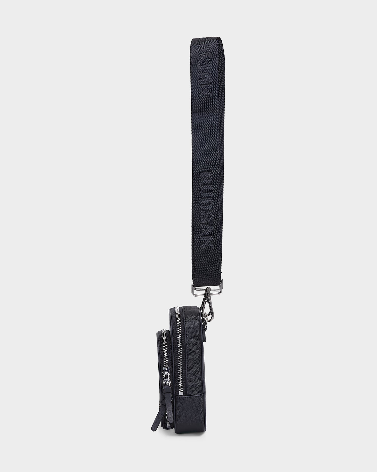 Side view of the JIMIE - L324505 Black, a small Rudsak crossbody bag with two zippered compartments and a wide black adjustable strap featuring raised RUDSAK lettering, shown against a plain light background.