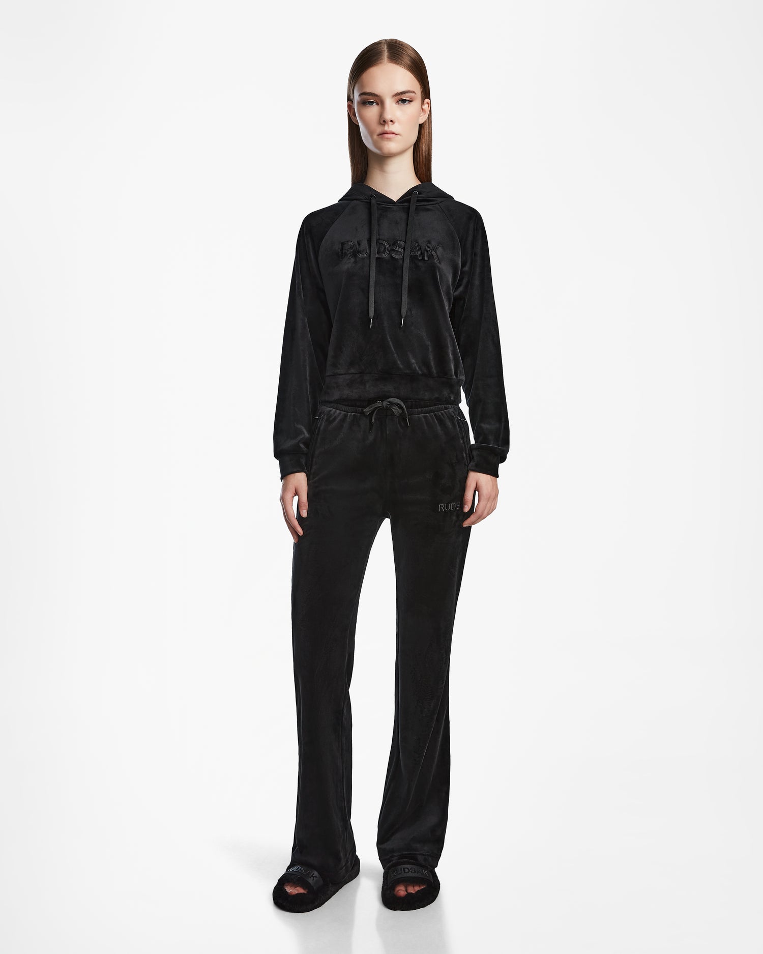 A woman stands facing forward in the Rudsak JODIE - L523501 Black velour tracksuit with a hooded sweatshirt and straight-leg pull-on pants, paired with black slides, set against a plain white background.