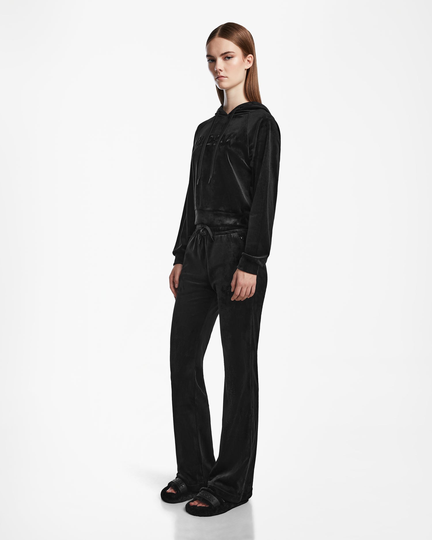 A woman wears the Rudsak JODIE - L523501 Black velour tracksuit, featuring a hooded sweatshirt and matching straight-leg elastic waist pants, against a plain white background, paired with black sandals.