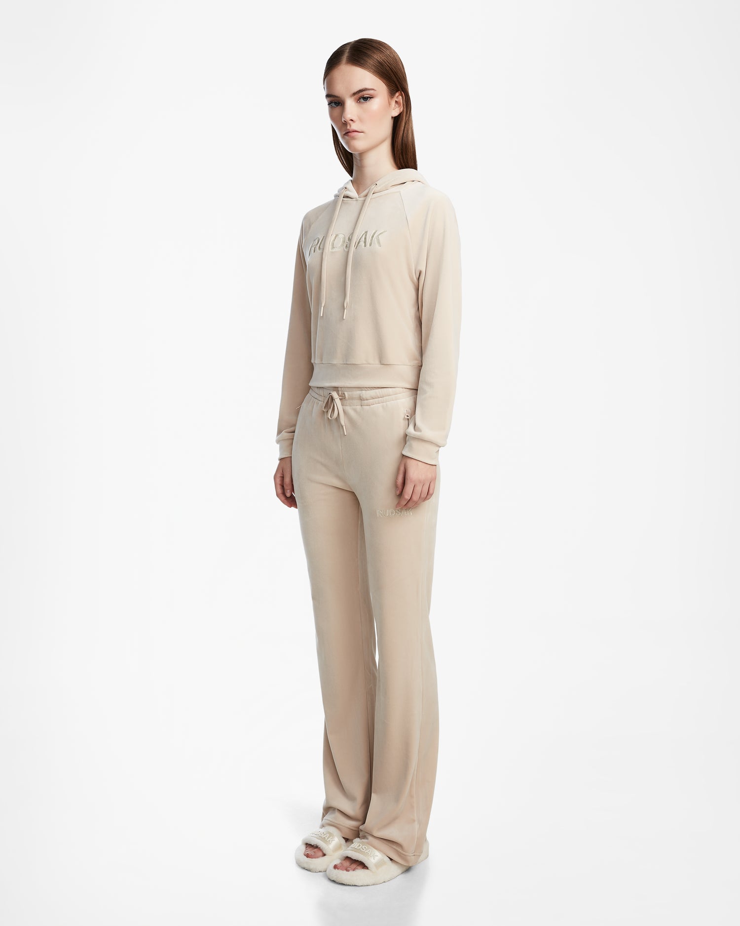 A woman wears the Rudsak JODIE - L523501 Cream velour tracksuit, featuring a cropped hoodie and elastic-waist velvet pull-on pants, paired with white fluffy slide sandals, set against a plain white background.