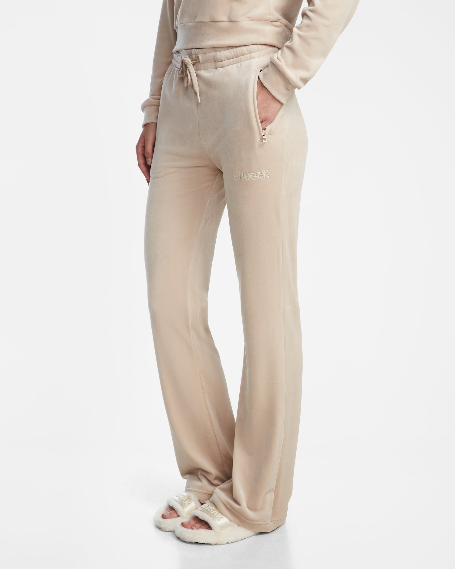 A person wearing Rudsak JODIE - L523501 Cream women’s velvet pull-on pants with an elastic waistband and zippered pockets, paired with fuzzy slippers, cropped from the shoulders down against a white background.