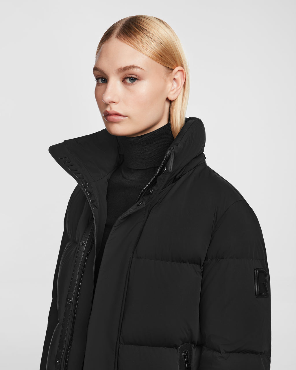 A woman with straight blonde hair wears a black turtleneck and the Rudsak JOON - 8124728 Black RDS-certified down puffer jacket with a high collar, posing against a plain light gray background.