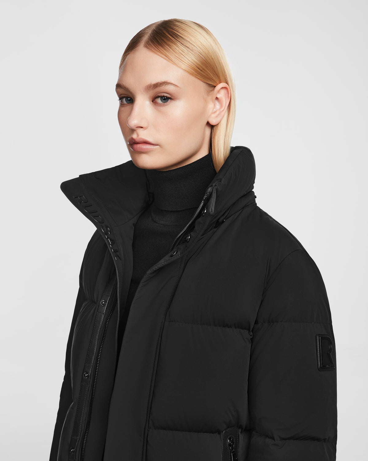 A woman with straight blonde hair wears a black turtleneck and the Rudsak JOON - 8124728 Black RDS-certified down puffer jacket with a high collar, posing against a plain light gray background.