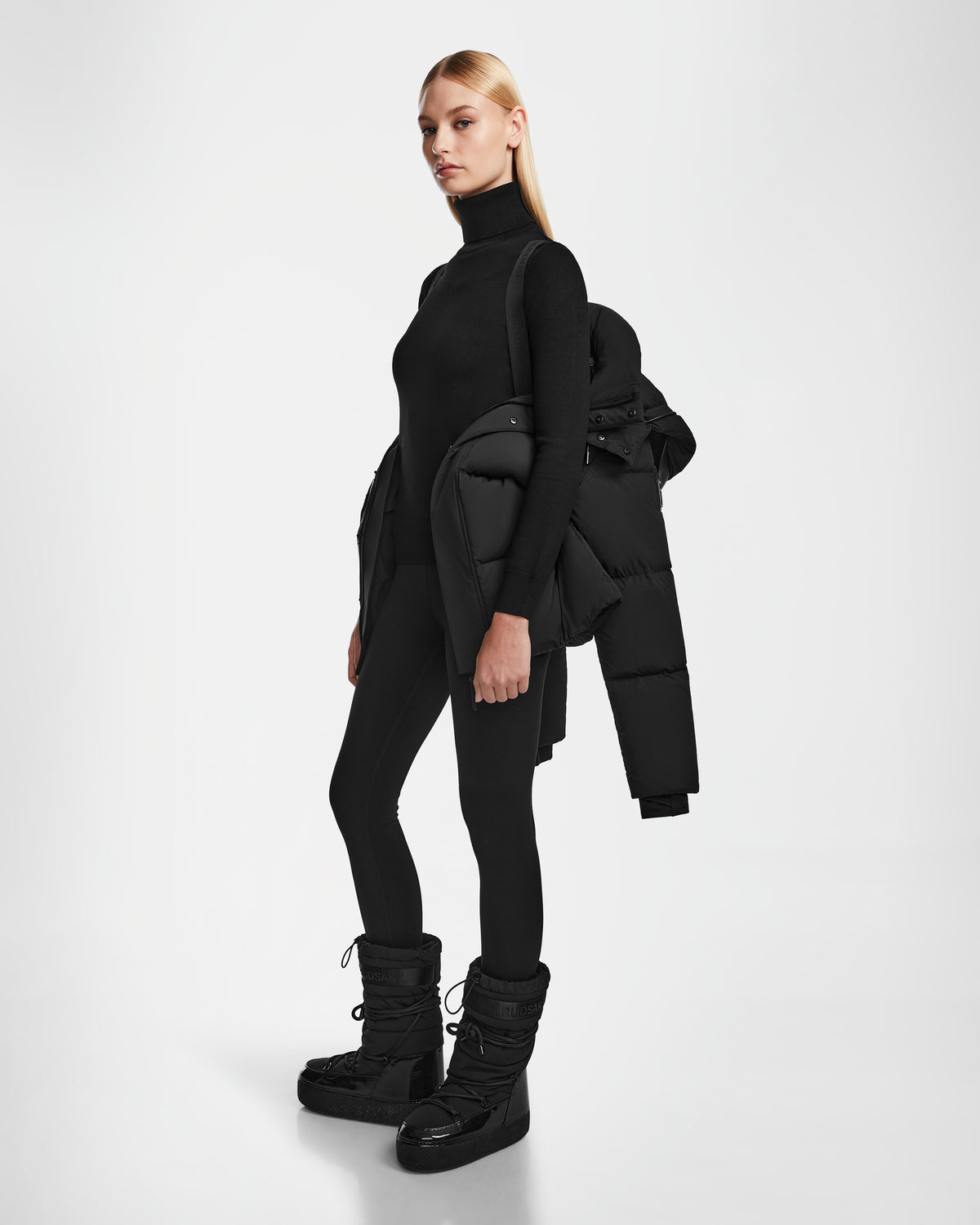 A woman with straight blonde hair and a serious expression stands against a white background, wearing Rudsaks JOON - 8124728 Black down puffer jacket draped over one shoulder, along with a black turtleneck, leggings, and puffer boots.