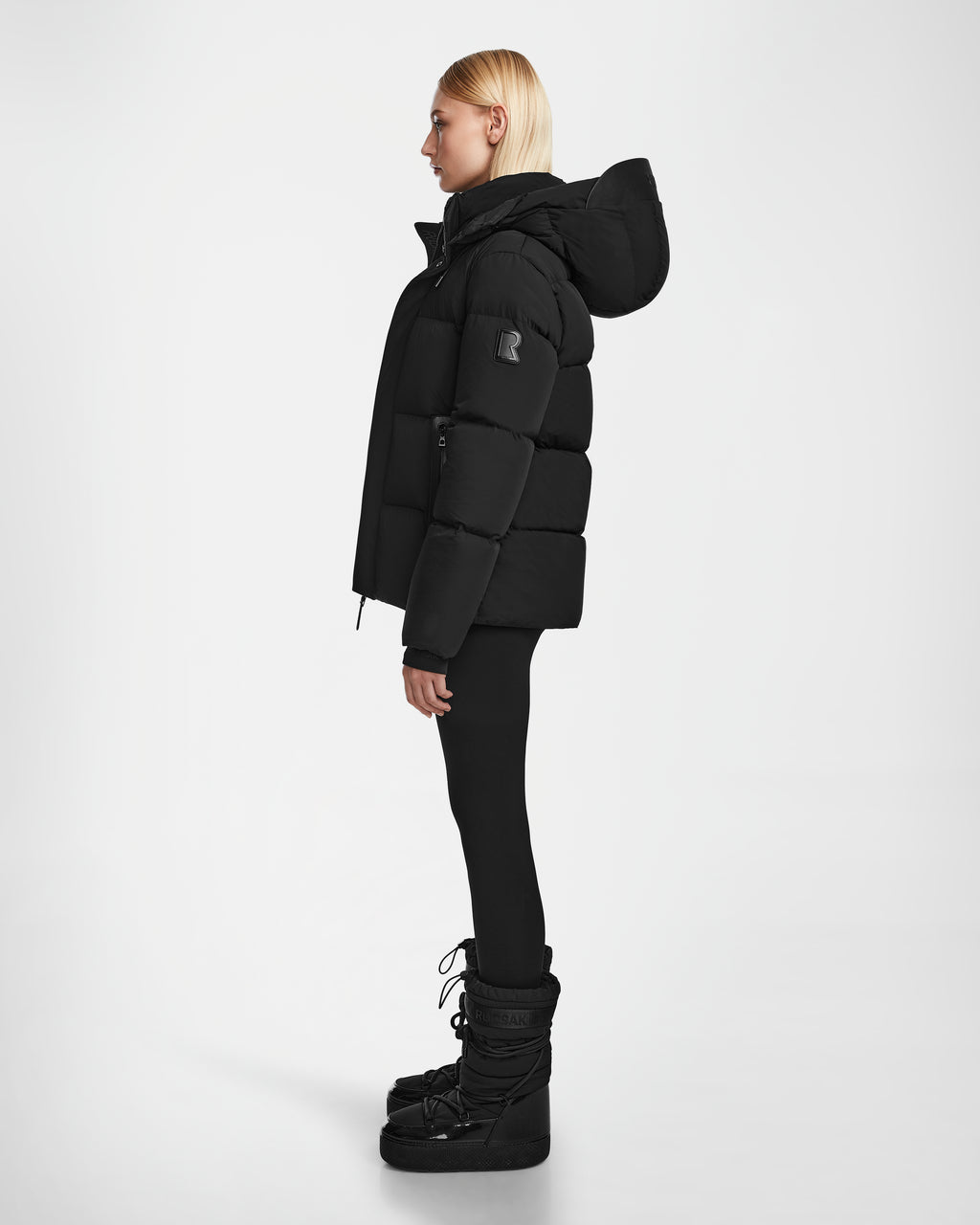 A woman with straight blonde hair stands in profile against a white background, wearing the Rudsak JOON - 8124728 Black puffer jacket, black leggings, and black snow boots.