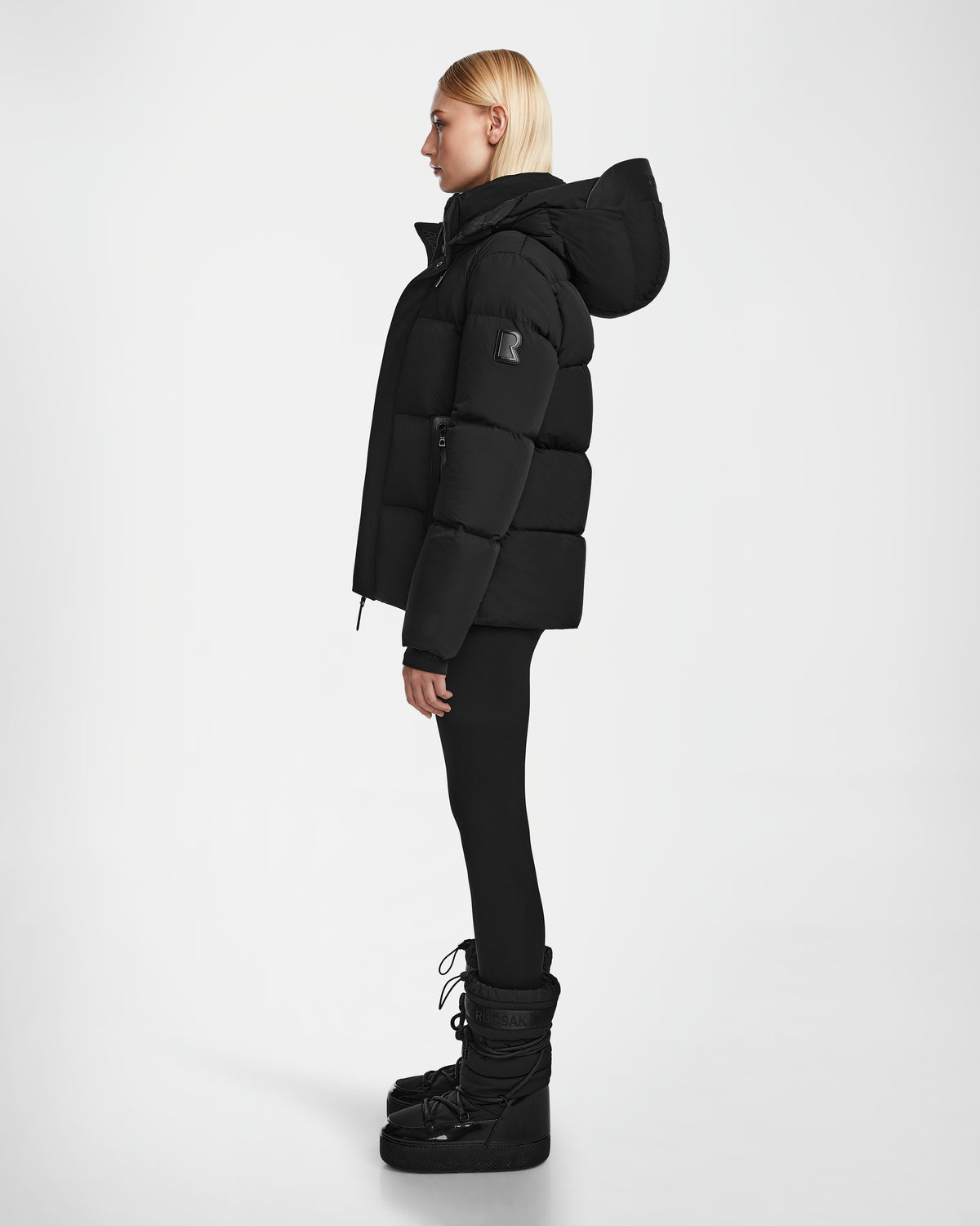 A woman with straight blonde hair stands in profile against a white background, wearing the Rudsak JOON - 8124728 Black puffer jacket, black leggings, and black snow boots.