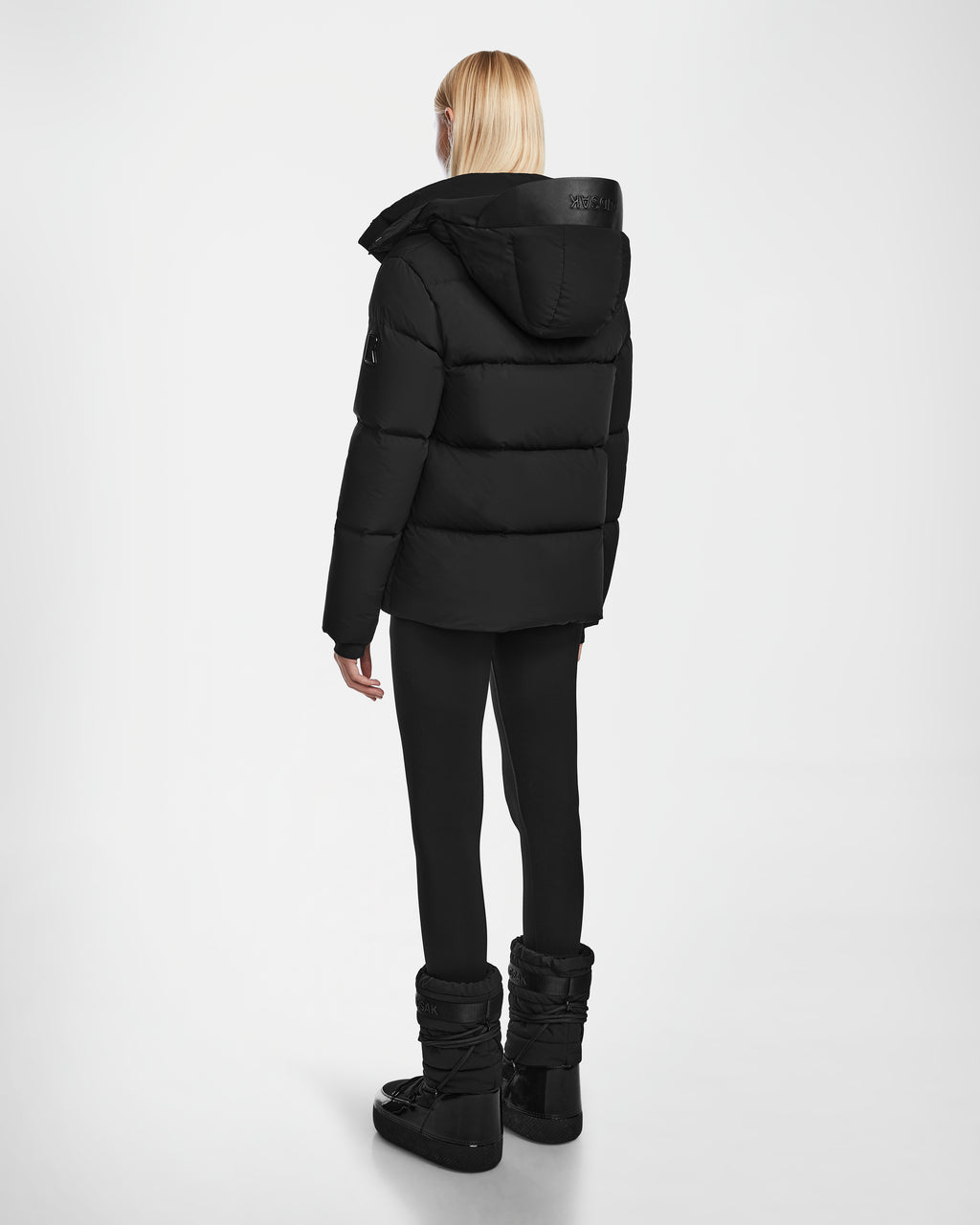 A person with straight blonde hair faces away, wearing the JOON - 8124728 Black RDS-certified down puffer jacket with hood by Rudsak, paired with black leggings and winter boots against a plain white background.