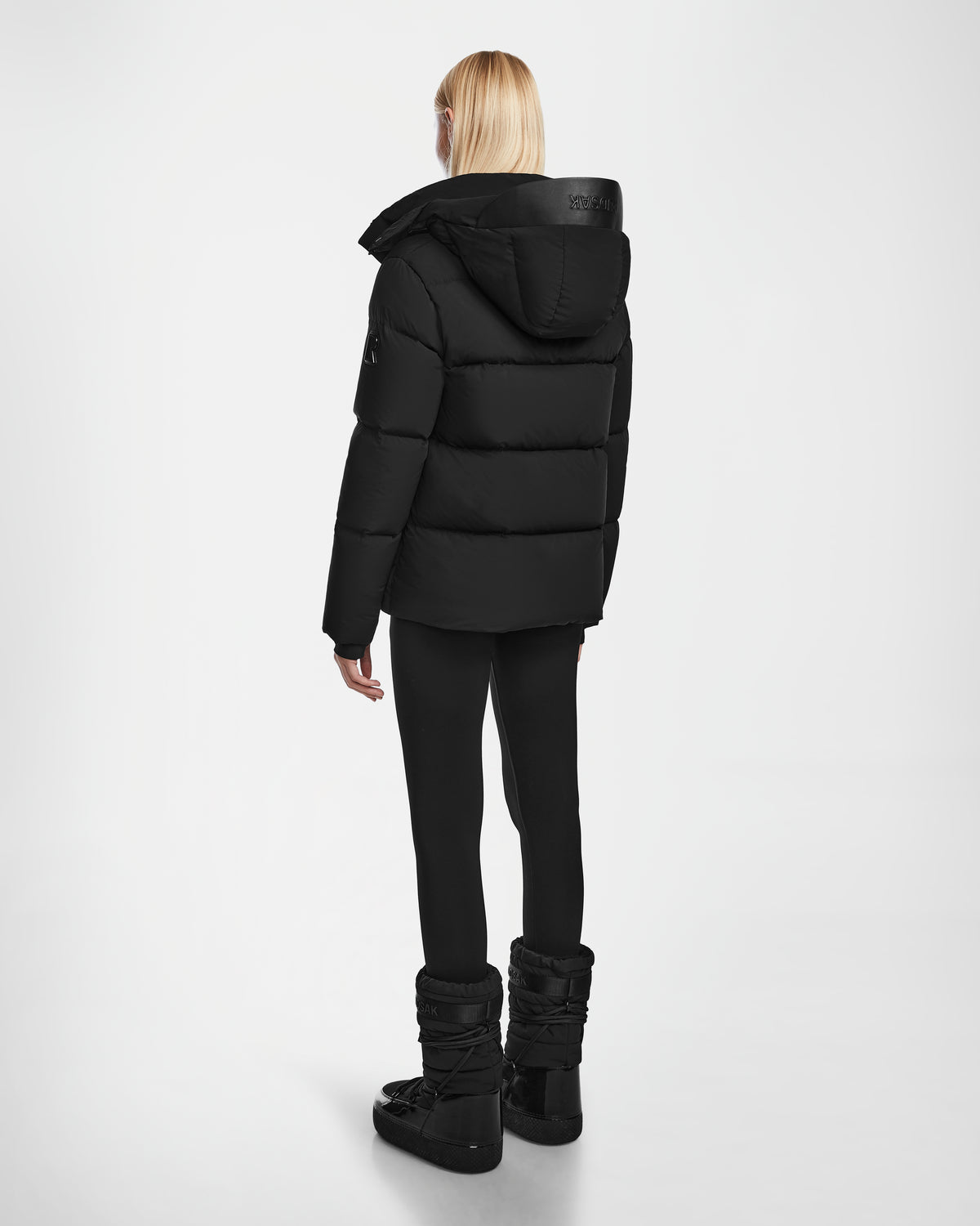 A person with straight blonde hair faces away, wearing the JOON - 8124728 Black RDS-certified down puffer jacket with hood by Rudsak, paired with black leggings and winter boots against a plain white background.