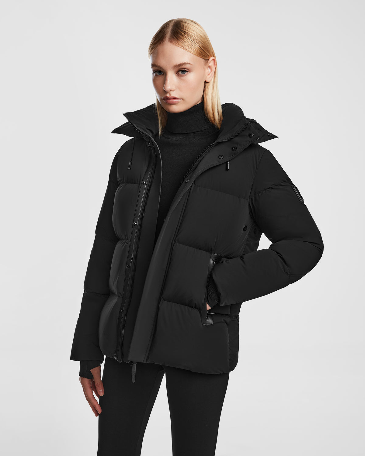 A woman with straight blonde hair models the Rudsak JOON - 8124728 Black down puffer jacket over a black turtleneck and pants, standing against a plain white background with a neutral expression.