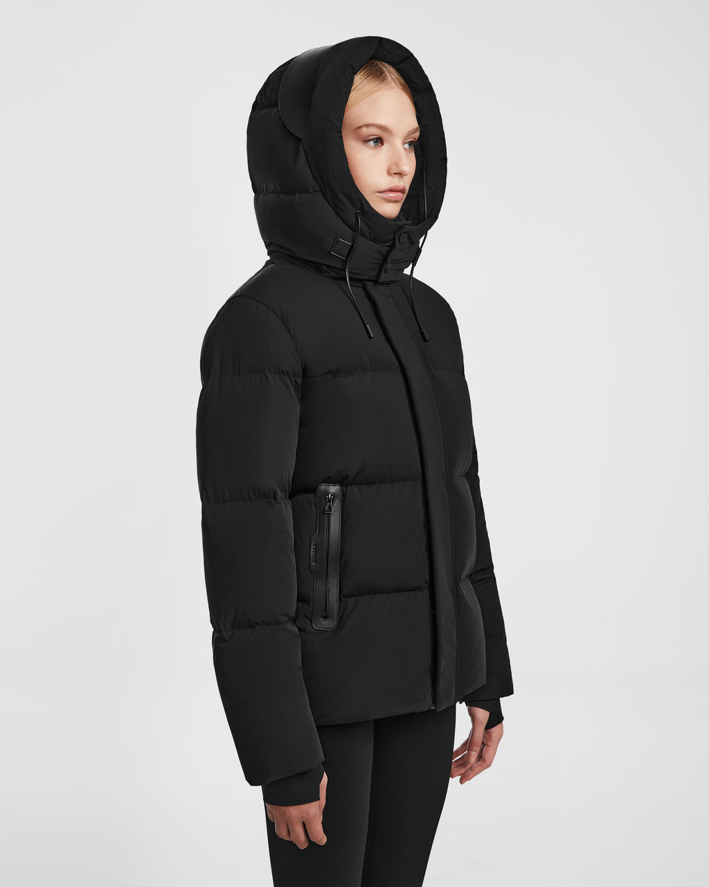 A person wearing the Rudsak JOON - 8124728 Black puffer jacket with a hood and zippered pockets stands against a plain white background, facing slightly to the side.