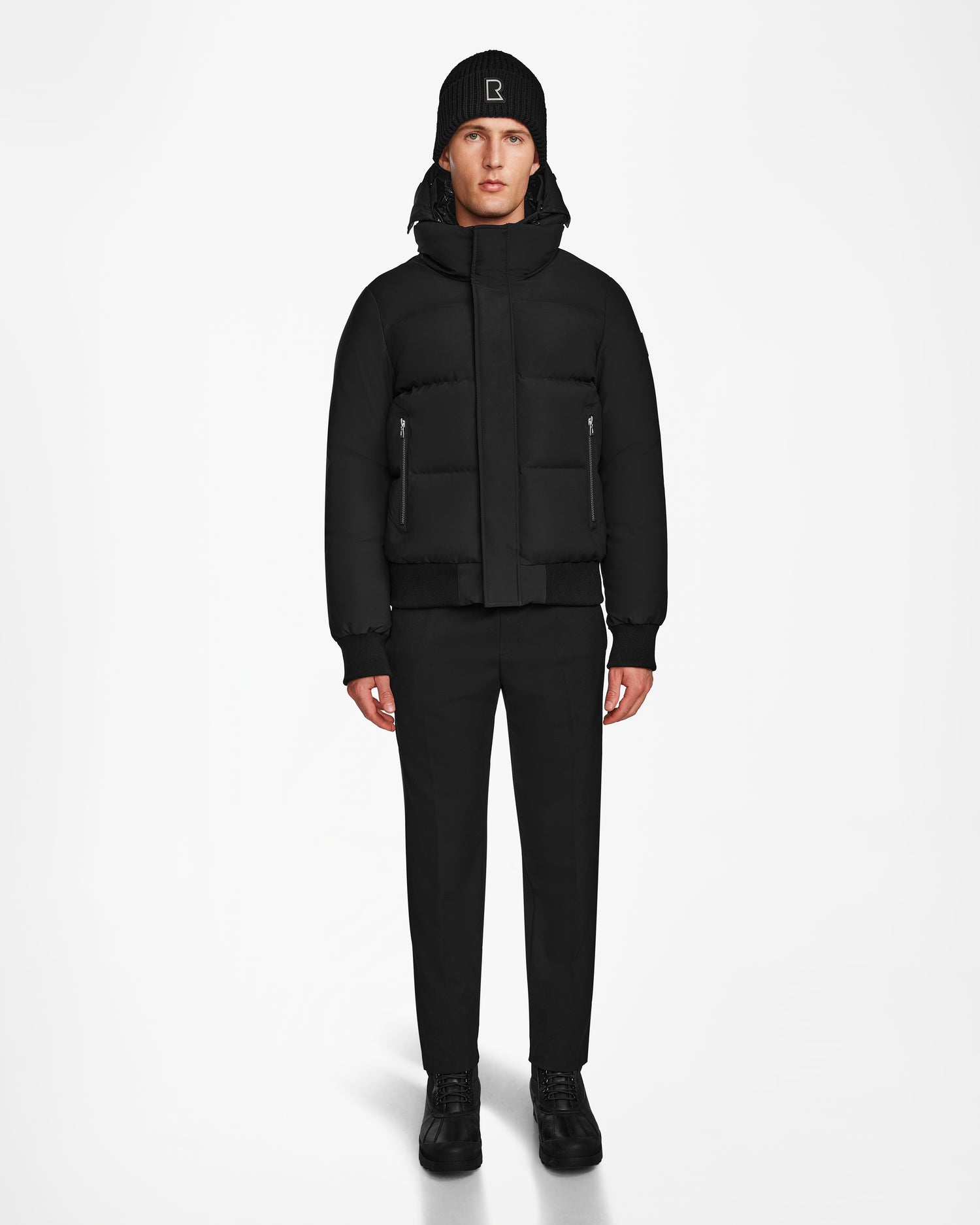 A man faces forward, wearing a Rudsak JORDY - 9123513 Black M unisex down puffer jacket, with black pants, black shoes, and a black beanie featuring a white logo, set against a plain white background.