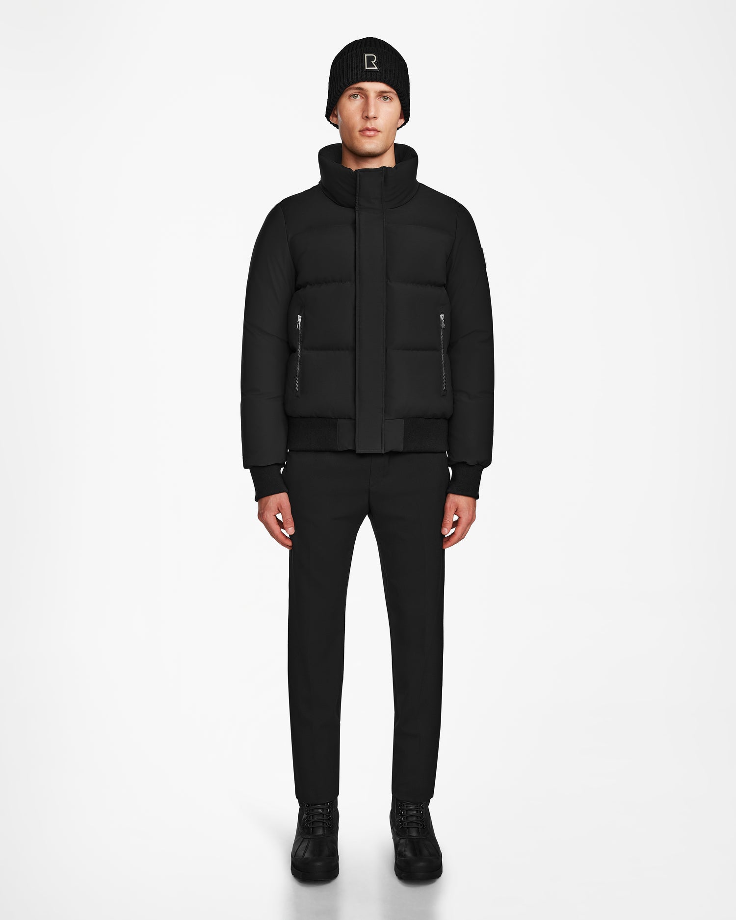 A person stands facing forward, wearing a black Rudsak JORDY - 9123513 M down puffer jacket with zippered pockets, paired with a black beanie, black pants, and black shoes against a plain white background.