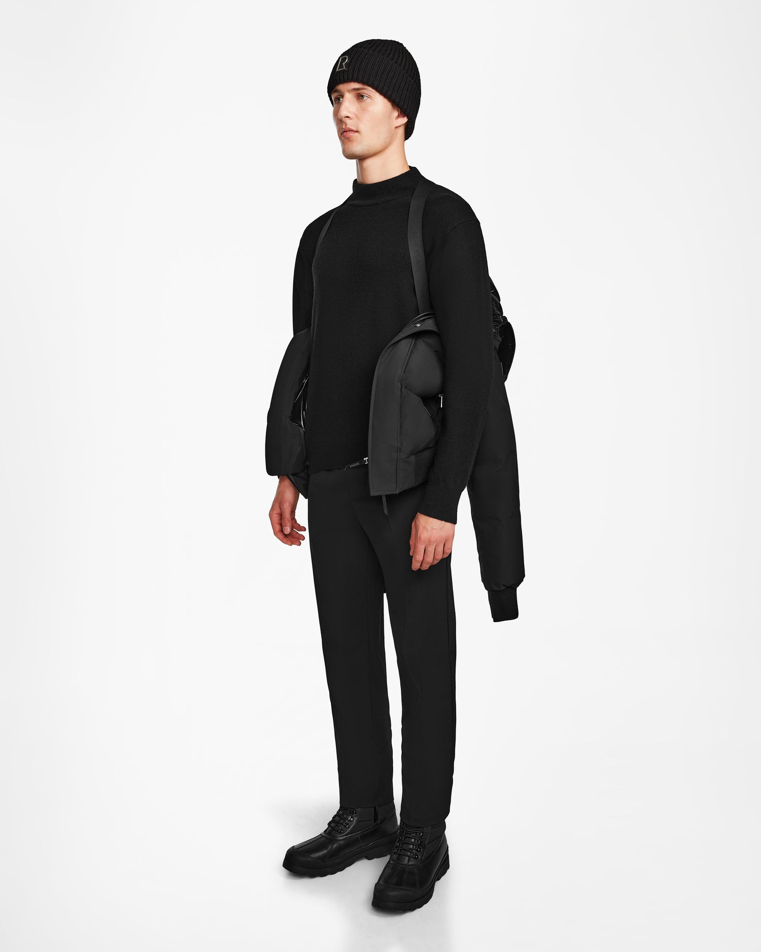 A man stands against a white background in black attire, with a Rudsak JORDY - 9123513 Black M recycled stormshell jacket draped over one shoulder and a black backpack on his back.