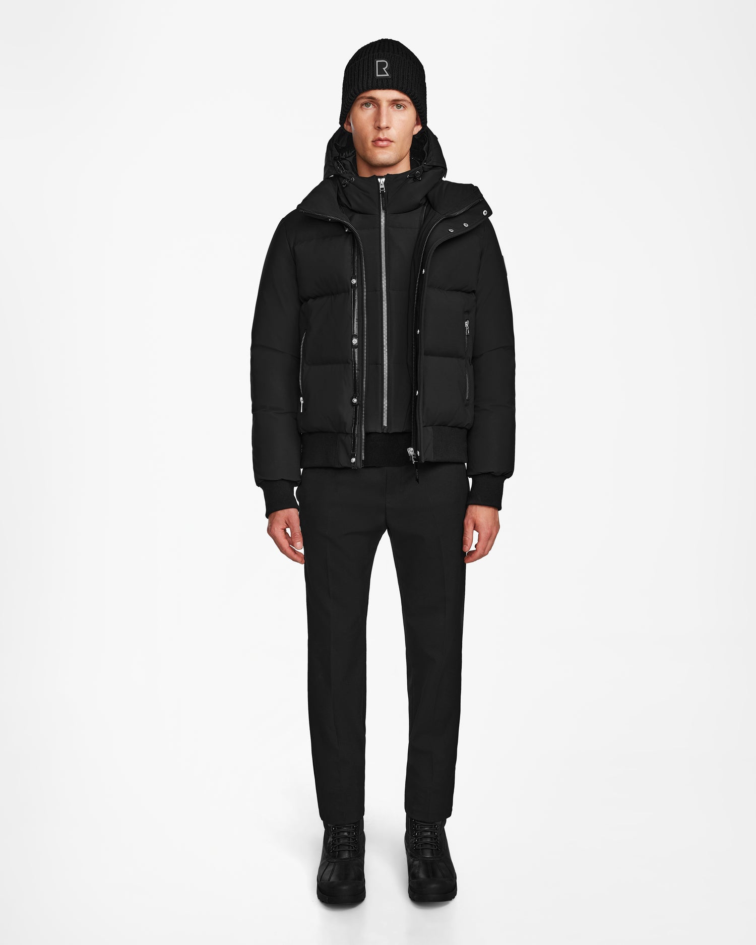 A person faces forward against a plain white background, wearing a black beanie, Rudsak JORDY - 9123513 Black M unisex down puffer jacket, zip-up hoodie, black pants, and black sneakers.