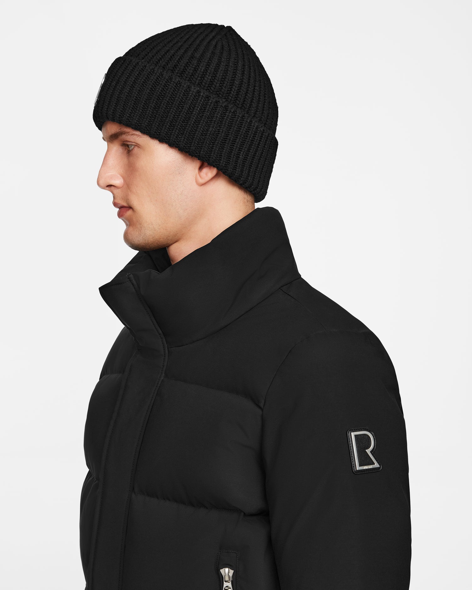 A young man in profile wears the Rudsak JORDY - 9123513 Black M unisex down puffer jacket with a high collar and an R logo on the sleeve, paired with a black knit beanie, against a plain white background.