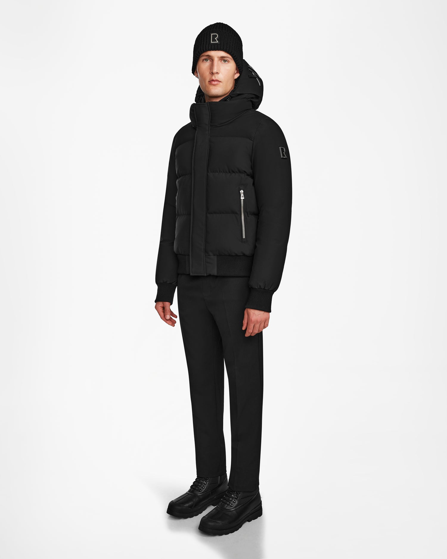 A man stands against a white background in a sleek all-black outfit, wearing the Rudsak JORDY - 9123513 Black M down puffer jacket featuring a front zipper and patch logo on the sleeve.
