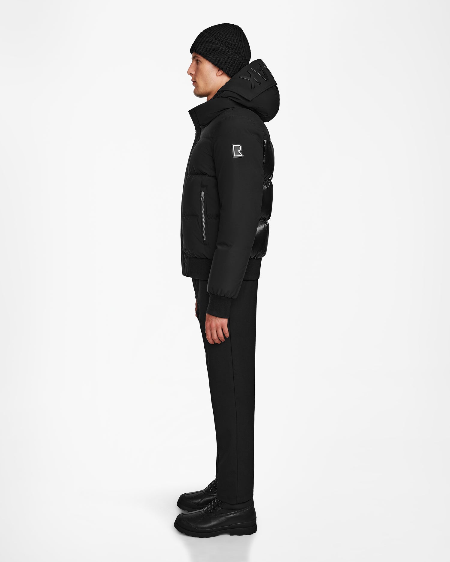 A person stands in profile against a white background, wearing a black beanie, Rudsak JORDY - 9123513 Black M down puffer jacket with visible sleeve logo, black pants, and black shoes.