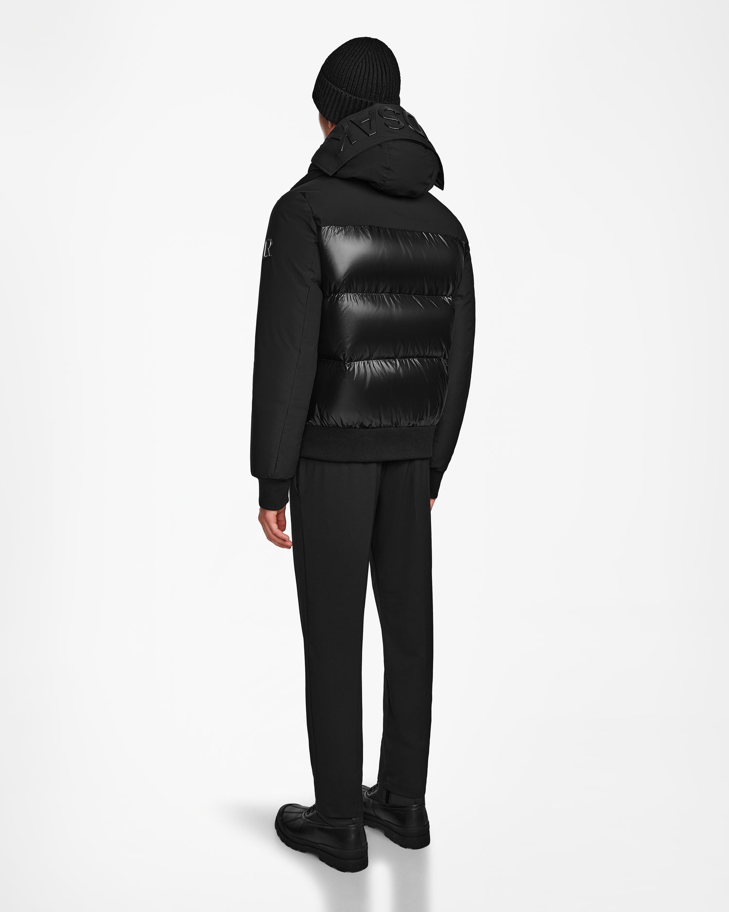 Dressed in all-black winter wear, including the Rudsak JORDY - 9123513 Black M down puffer jacket, beanie, pants, and boots, a person stands facing away against a plain white background.