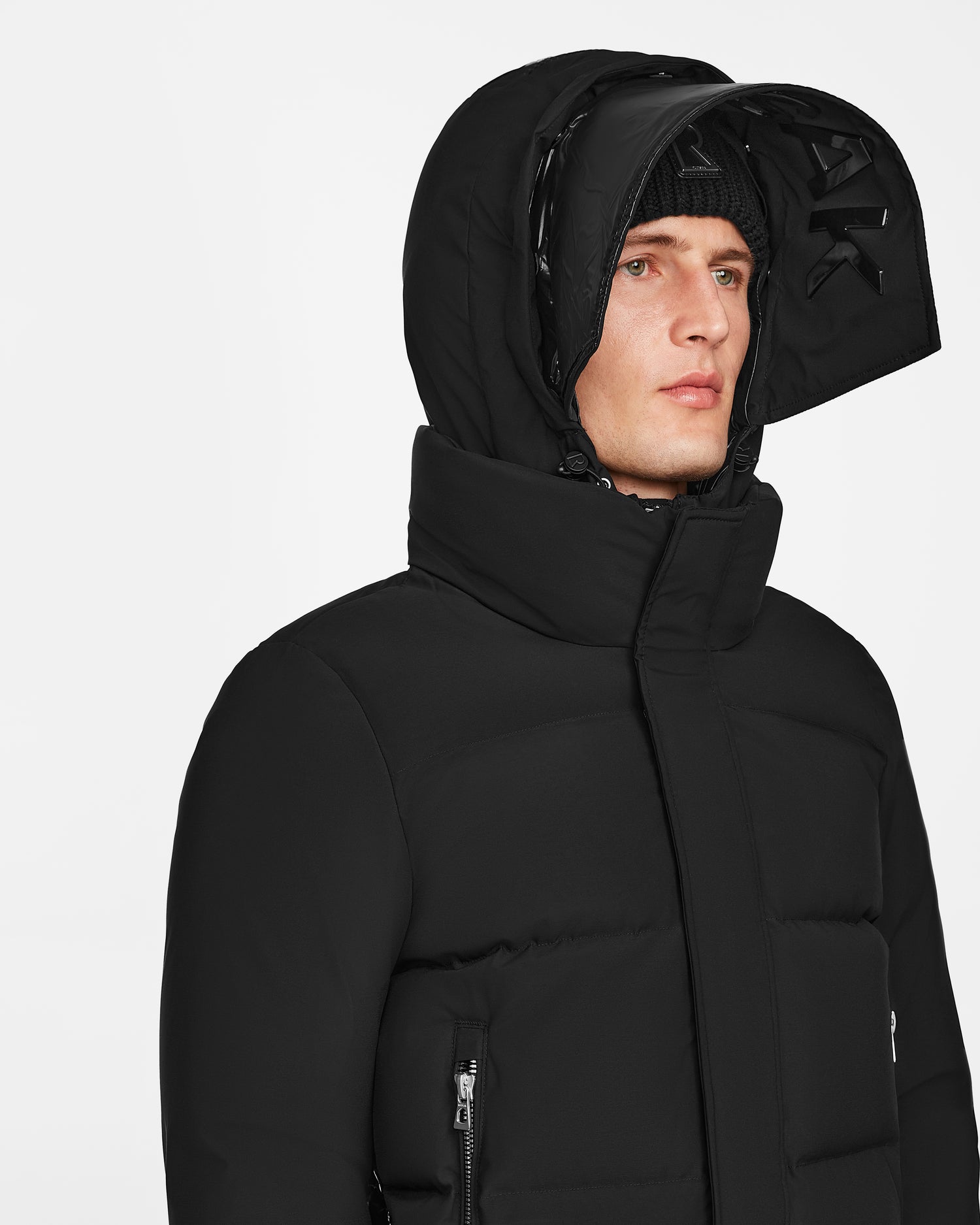 Someone wears a black Rudsak JORDY - 9123513 down puffer jacket, size M, with the hood up and partially unzipped, paired with a black beanie. The high-collar unisex stormshell jacket stands out against a plain light background.