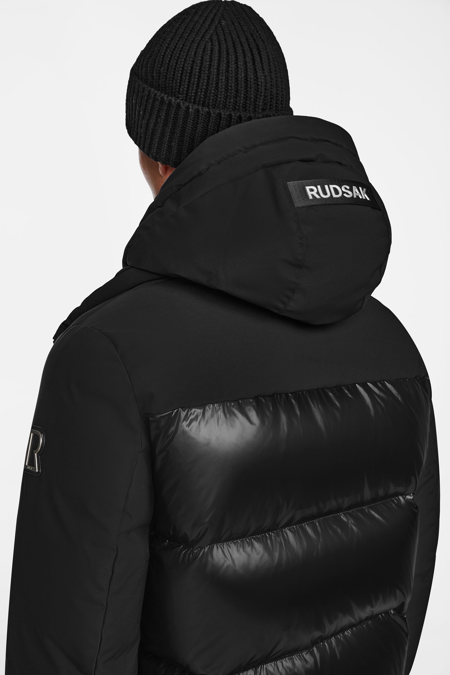 A person in a black Rudsak JORDY - 9123513 down puffer jacket and knit beanie, seen from behind with Rudsak on the hood, stands against a plain background.