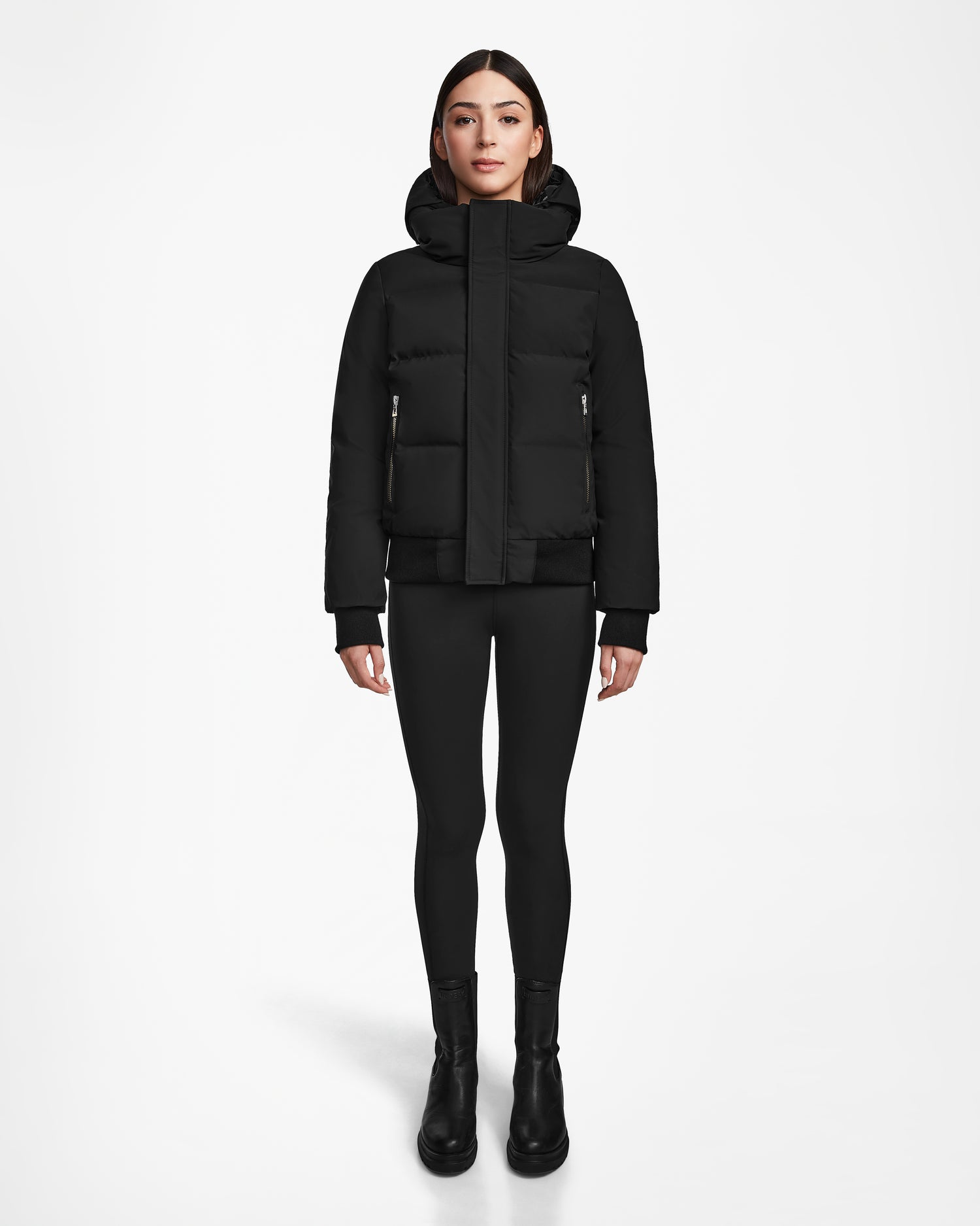 A woman stands facing forward against a plain white background, wearing the Rudsak JORDY - 9123513 Black W recycled stormshell jacket with a high collar, paired with black leggings and boots.