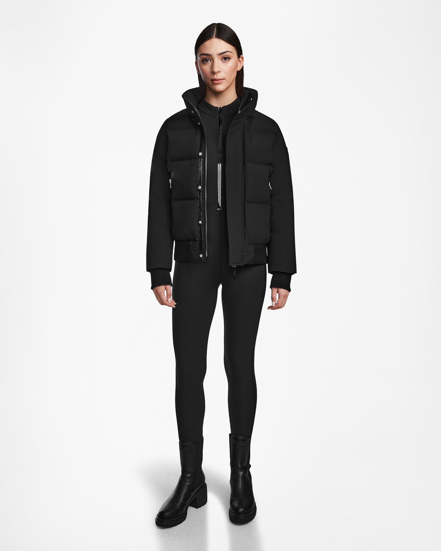 A woman faces forward in a plain white setting, wearing the sleek and modern Rudsak JORDY - 9123513 Black W winter jacket, paired with matching black leggings and boots.