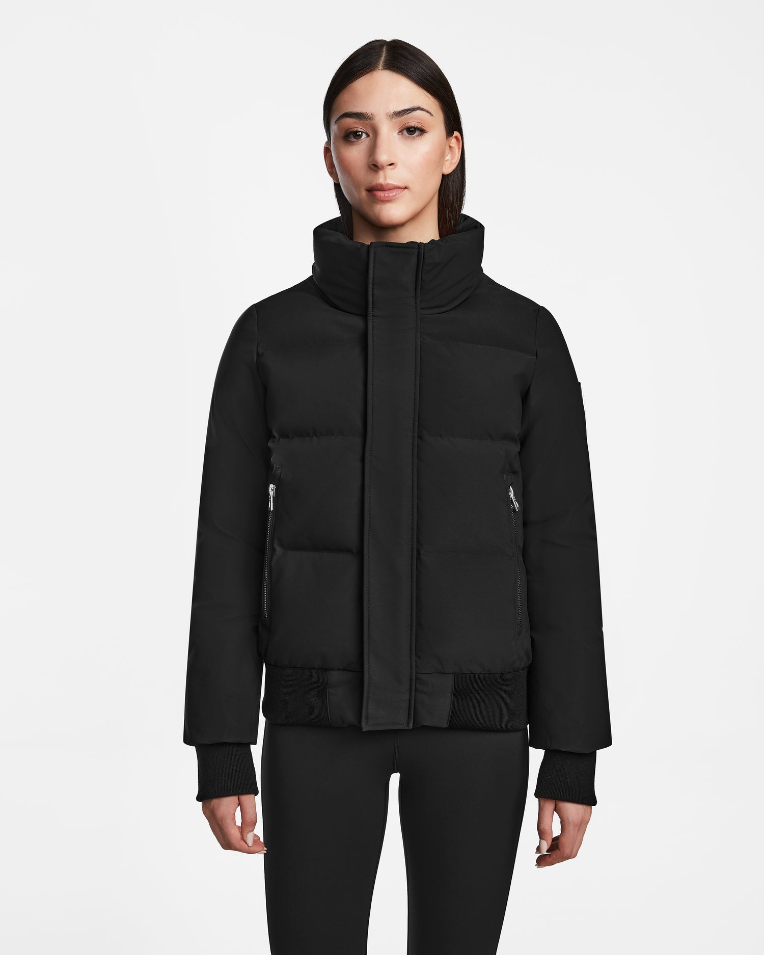 A woman with straight dark hair wears the Rudsak JORDY - 9123513 Black W unisex down puffer jacket and black leggings, standing facing forward against a plain white background.