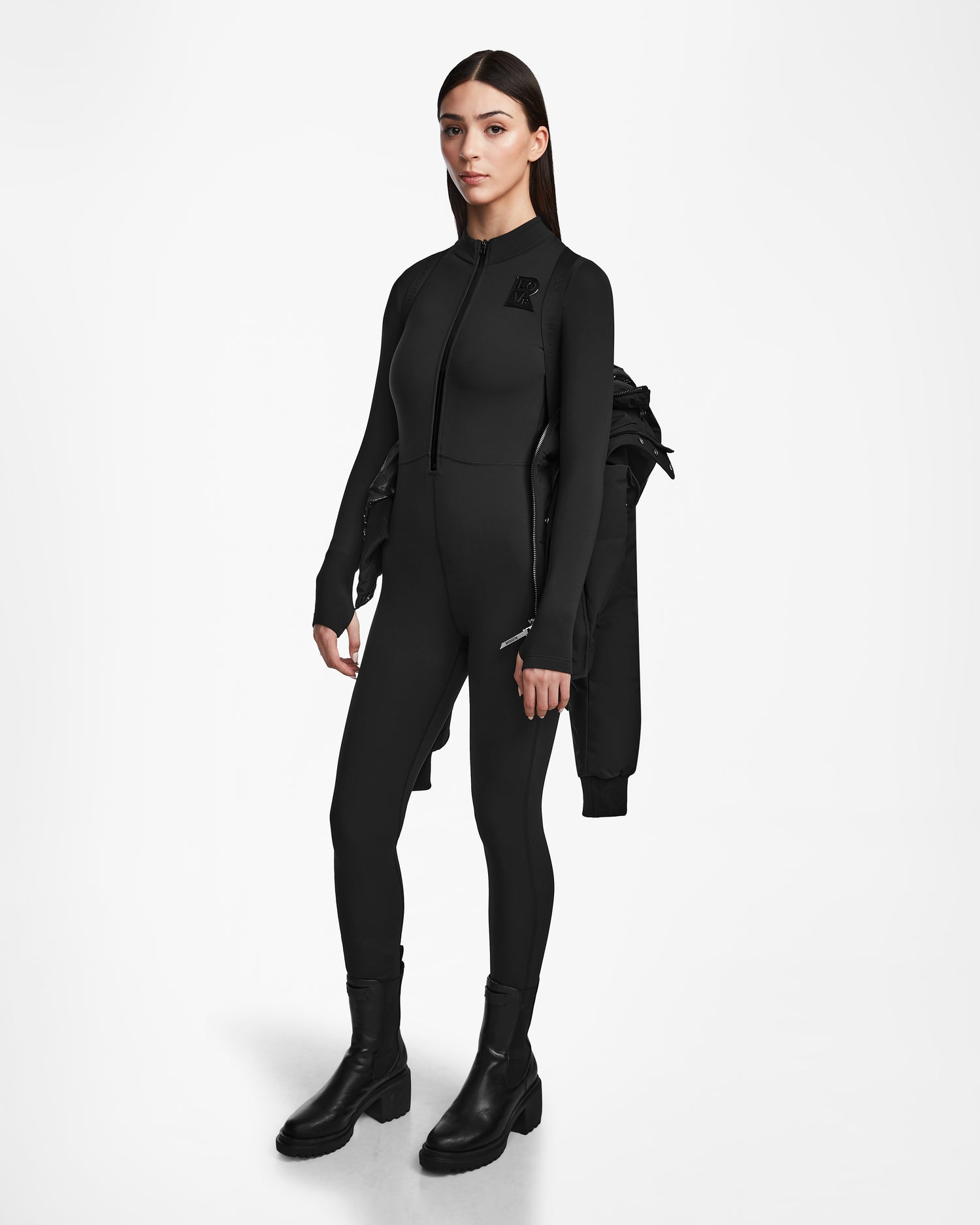 A woman with straight, loose hair and a neutral expression stands against a white backdrop, wearing the Rudsak JORDY - 9123513 Black W jumpsuit and black boots, with a recycled stormshell jacket draped over one shoulder.