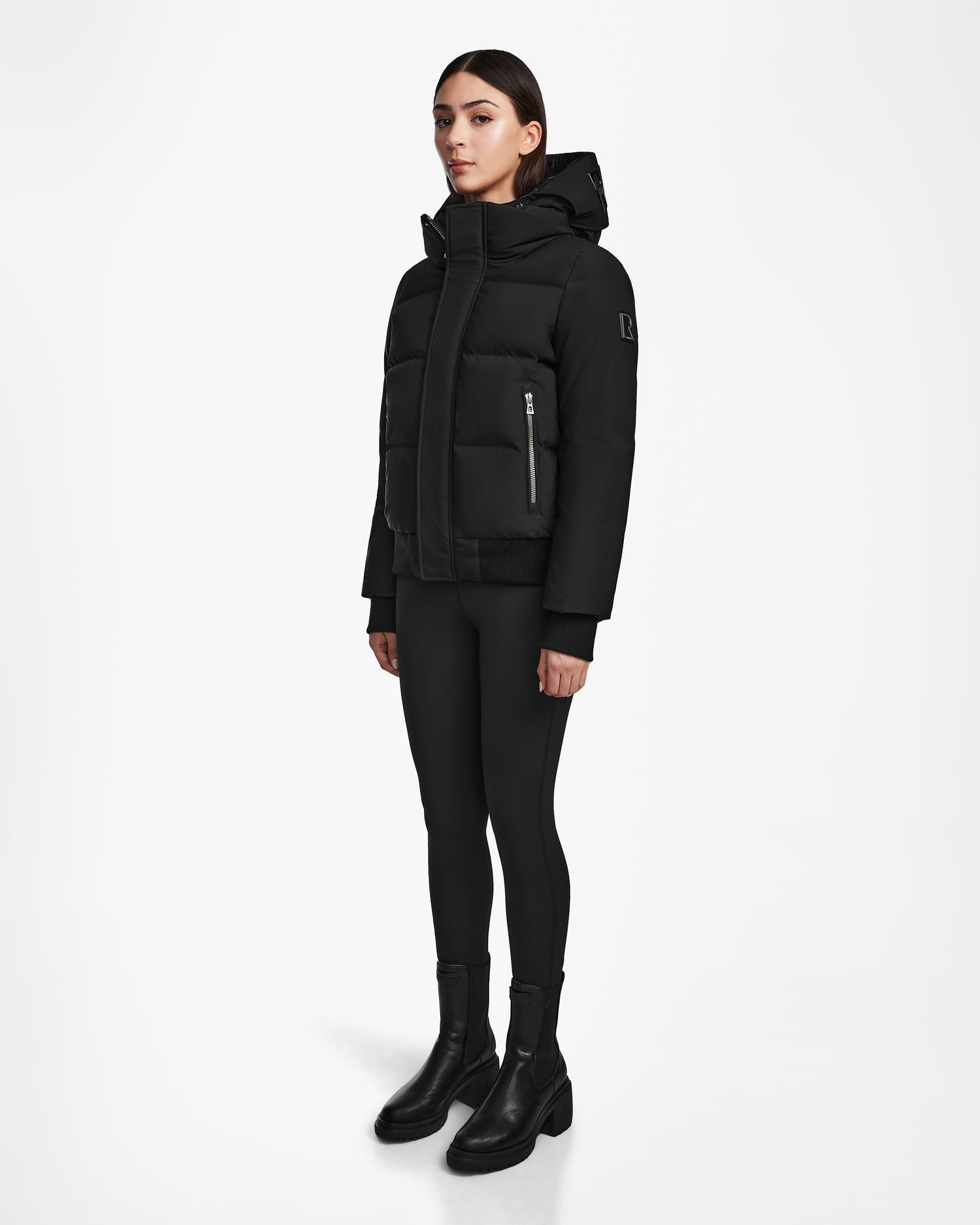 A woman stands against a white background wearing the Rudsak JORDY - 9123513 Black W jacket, paired with black leggings and ankle boots, facing forward with her arms relaxed at her sides.