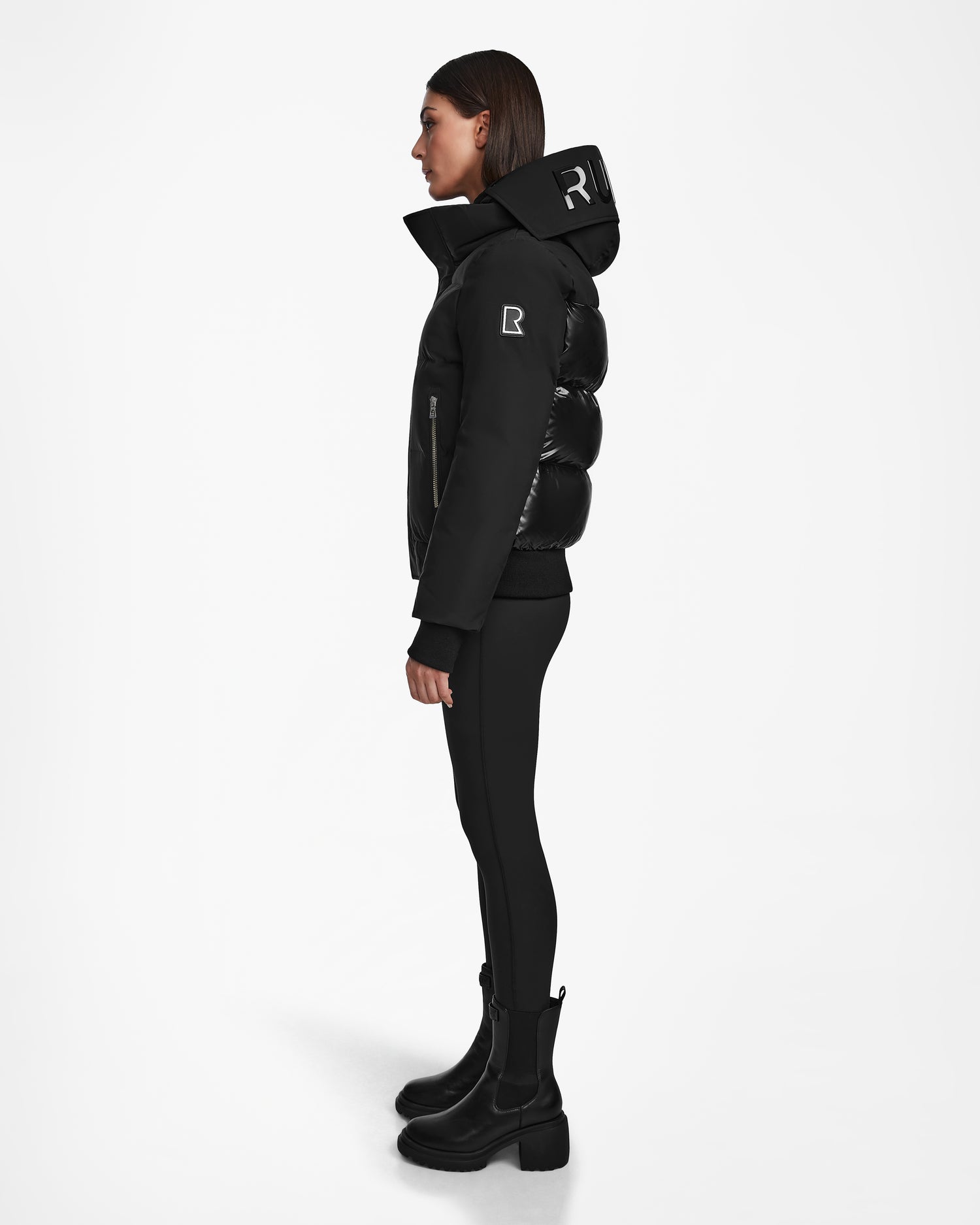 A woman in profile wears the Rudsak JORDY - 9123513 Black W recycled stormshell jacket with a raised collar, logo patches, and shiny quilted sections, styled with black leggings and heeled boots against a white background.