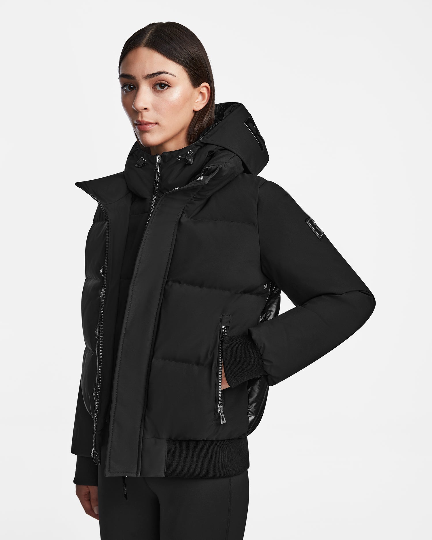 A woman with straight dark hair wears the Rudsak JORDY - 9123513 Black W down-filled jacket and black pants, standing against a plain white background.