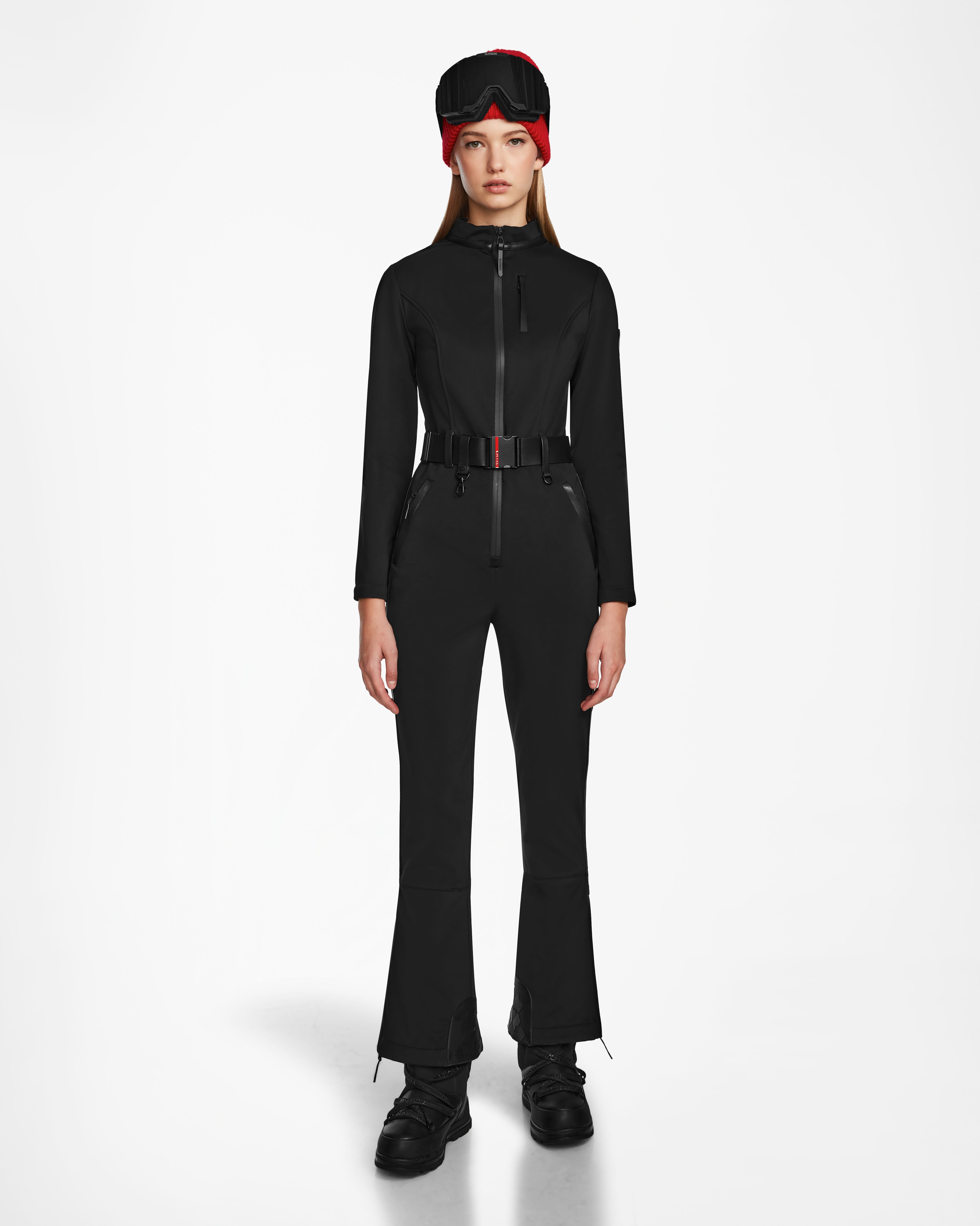A woman faces forward wearing the Rudsak JOSIE - 8124817 Black womens ski suit, styled with black boots and a red and black winter hat with goggles on top, set against a plain white background.