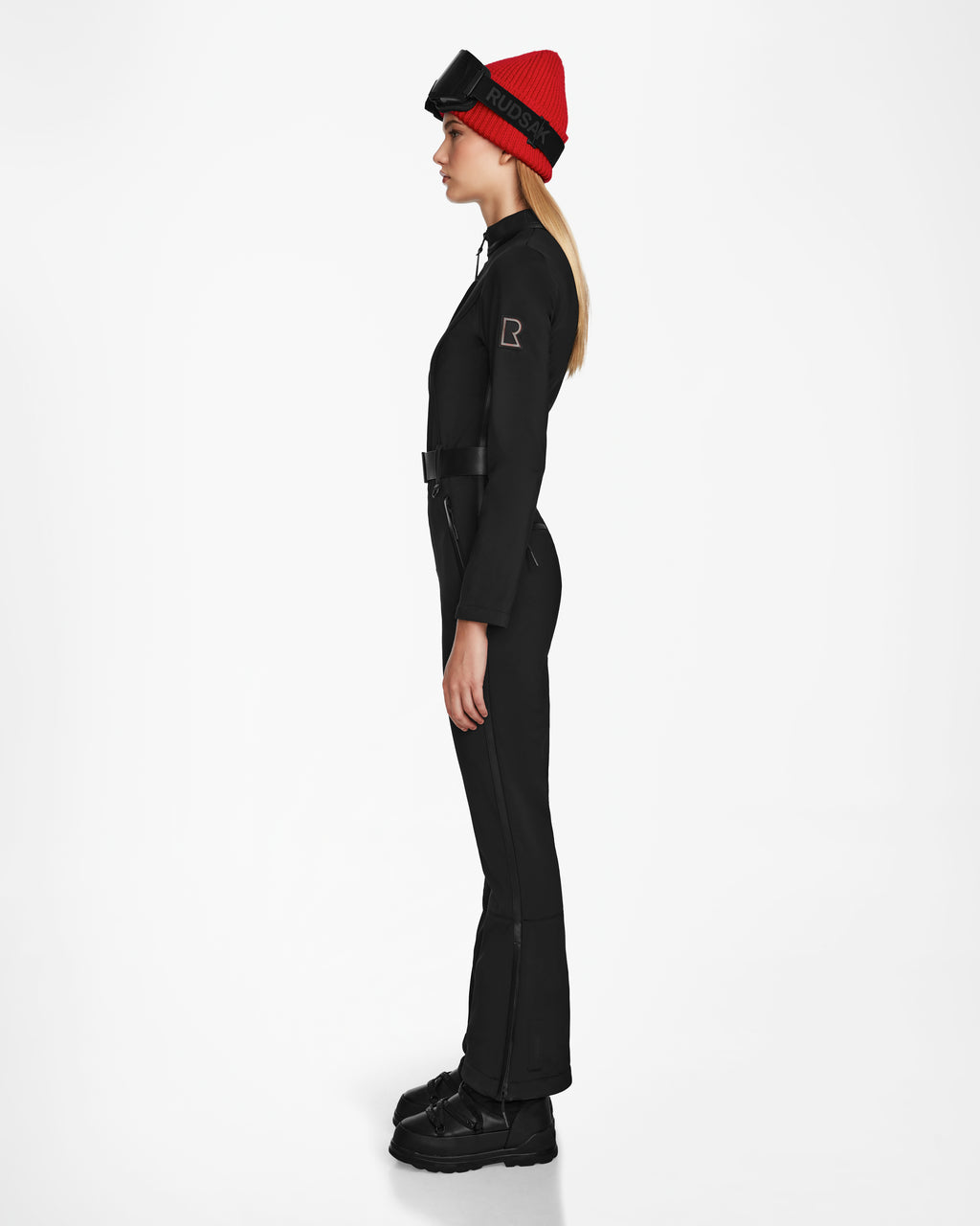 A woman stands in profile against a white background wearing Rudsak’s JOSIE - 8124817 Black women’s ski suit with technical stretch fabric, black goggles, a red beanie, and black boots.