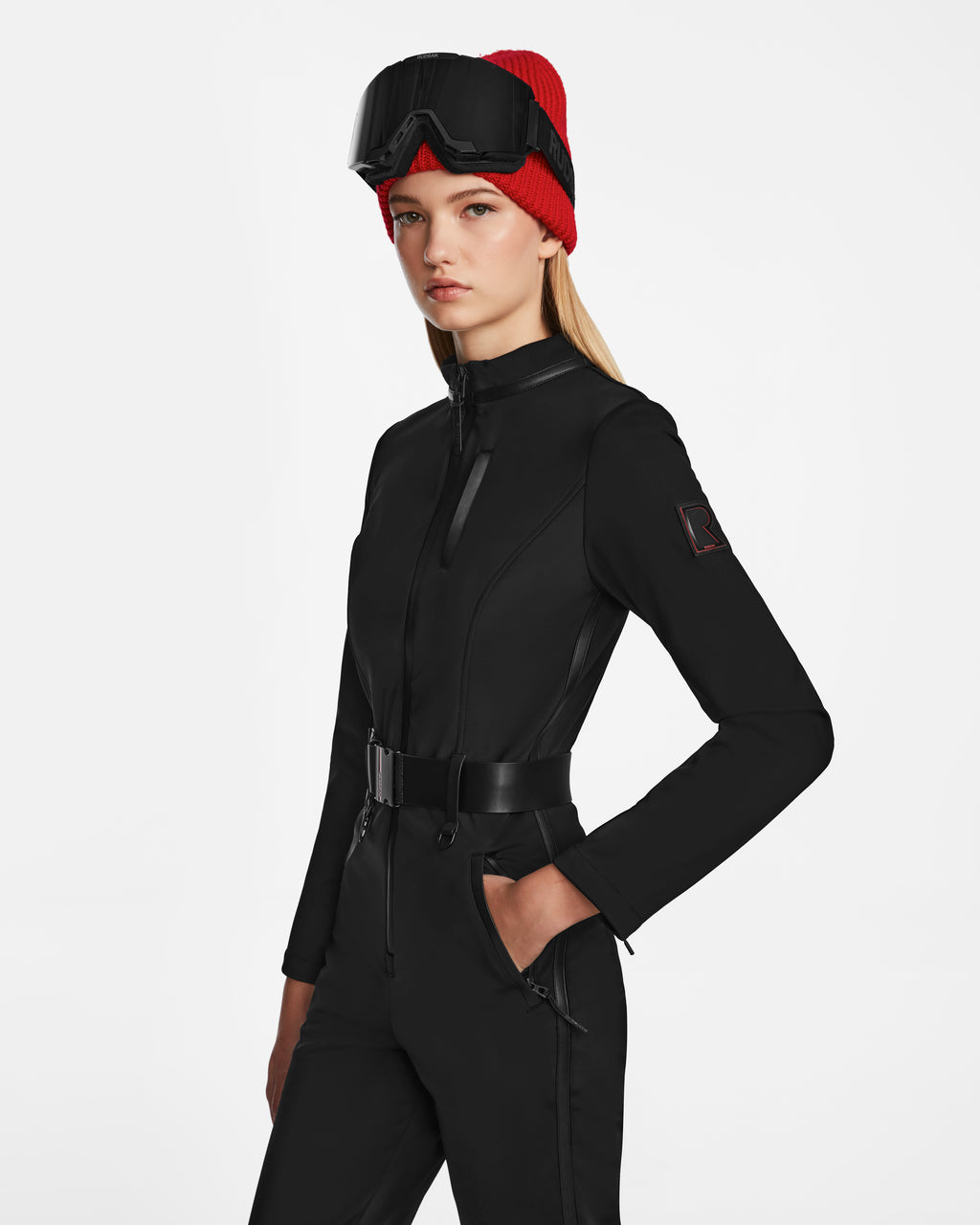 A woman wears the Rudsak JOSIE - 8124817 Black ski suit with technical stretch fabric, zip front, wide belt, and zipper pockets, paired with a red beanie and black ski goggles, standing against a plain white background.