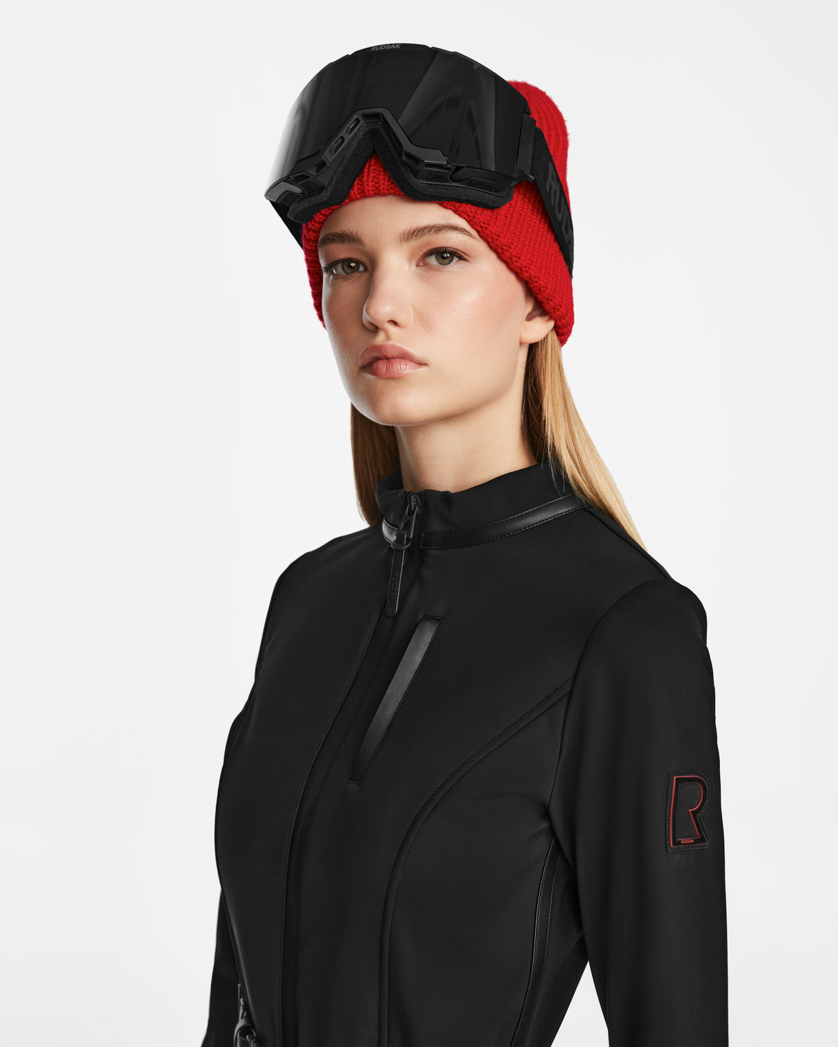 A young woman confidently poses against a white background, wearing the Rudsak JOSIE - 8124817 Black womens ski suit, paired with a red knit beanie and black ski goggles resting on her forehead.