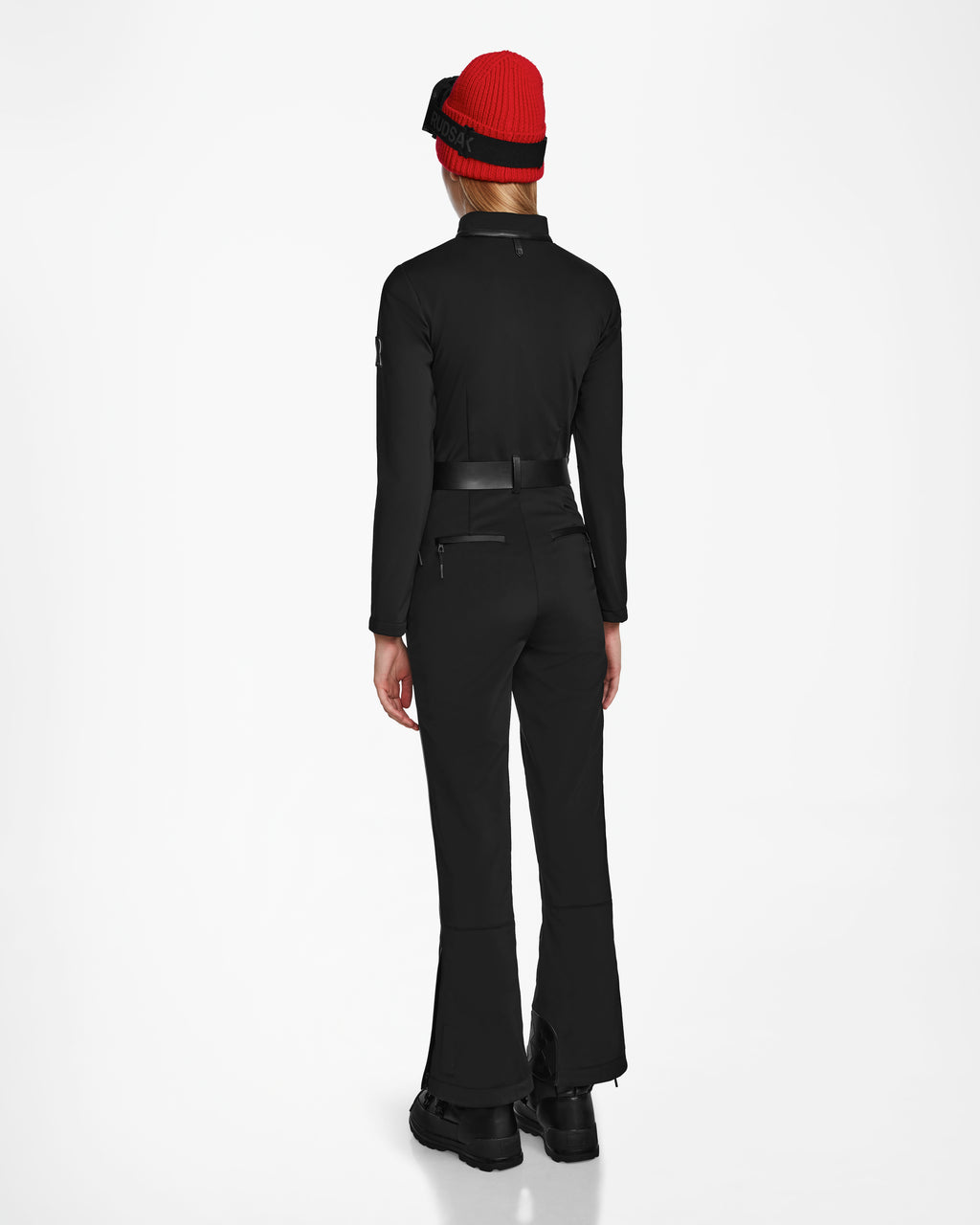 A person faces away from the camera, wearing the Rudsak JOSIE - 8124817 Black fitted jumpsuit, paired with black boots and a bright red knit hat with a black band. The background is plain white.