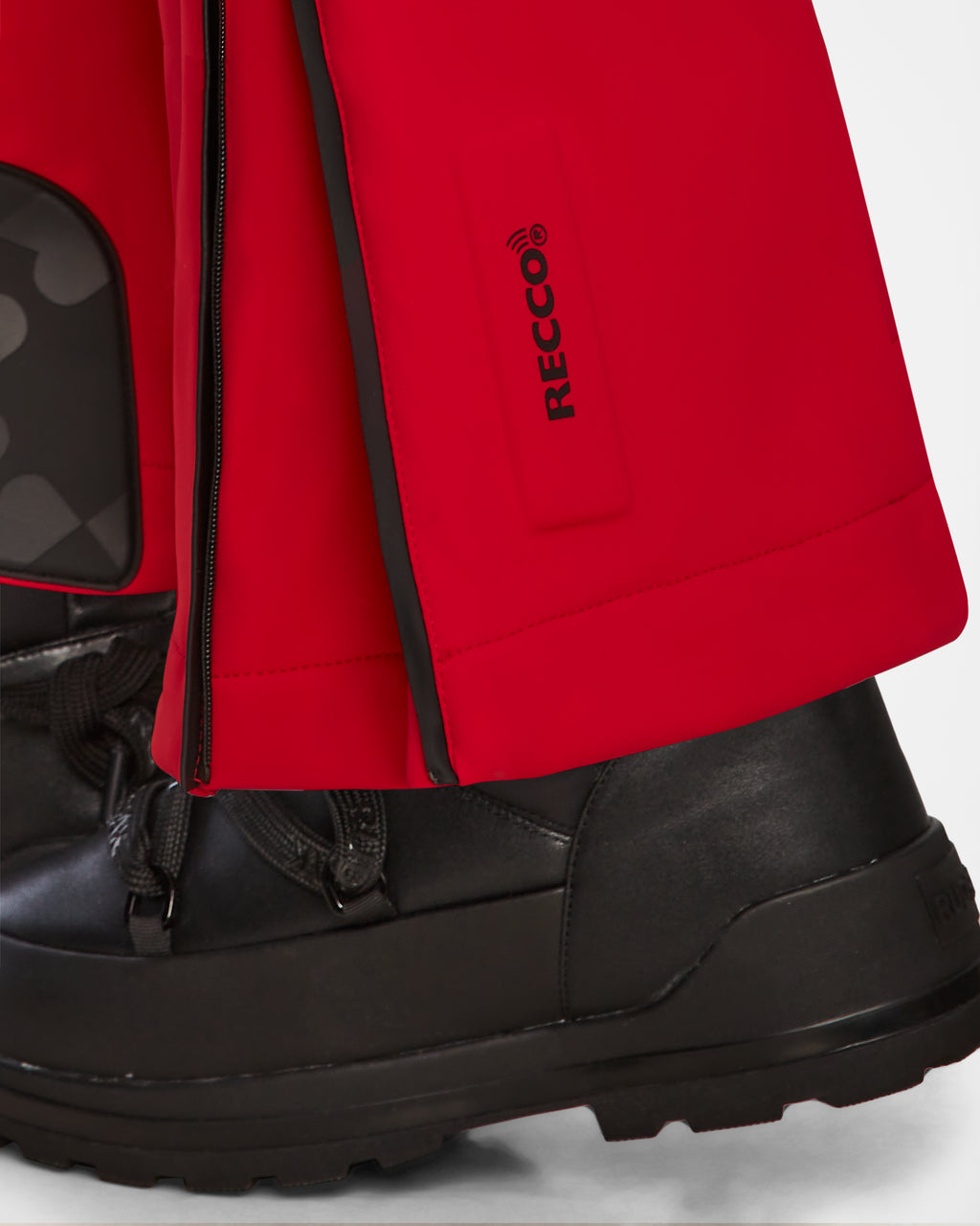 Close-up of a person wearing Rudsak’s JOSIE - 8124817 Flame Red women’s ski suit, featuring black boots, bright red pants with a RECCO rescue technology label, and a black zipper on the lower leg and foot.