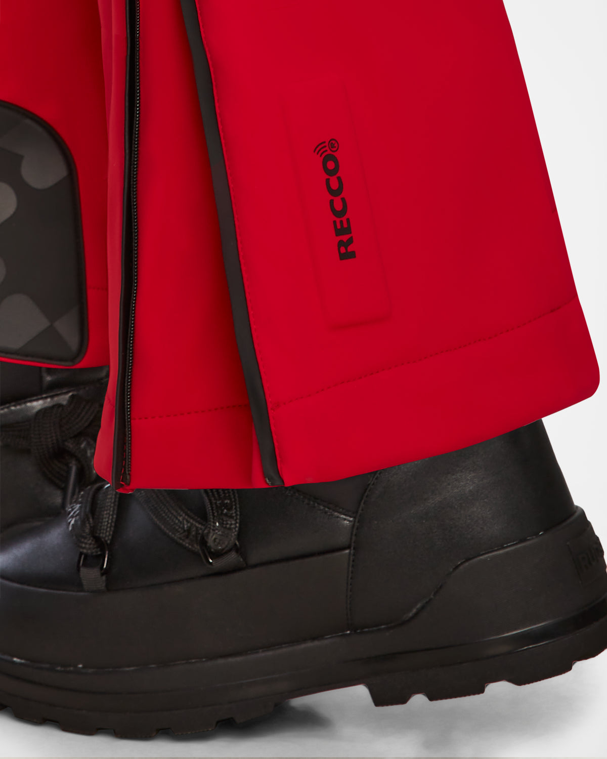 Close-up of a person wearing Rudsak’s JOSIE - 8124817 Flame Red women’s ski suit, featuring black boots, bright red pants with a RECCO rescue technology label, and a black zipper on the lower leg and foot.