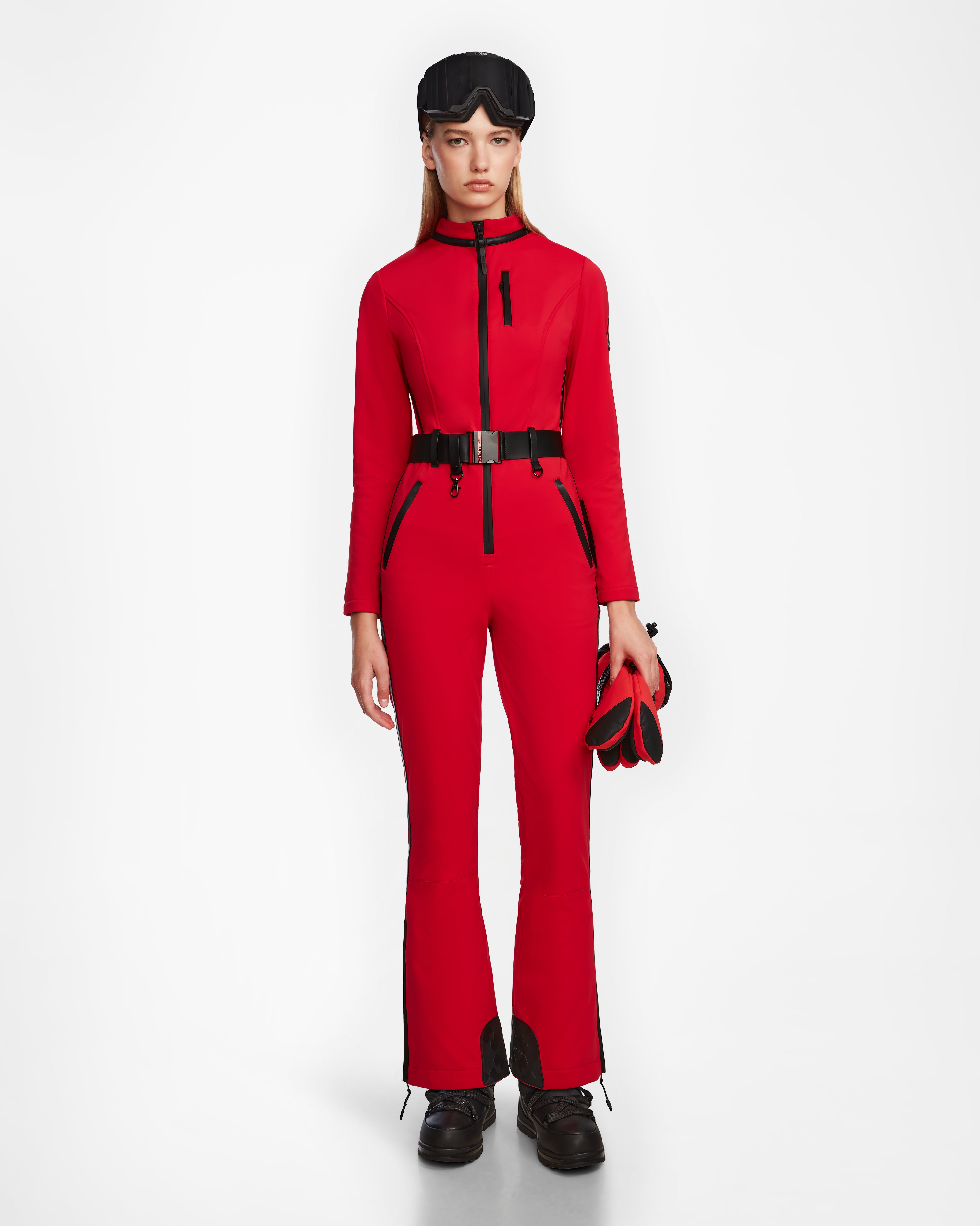 A woman faces forward wearing the Rudsak JOSIE - 8124817 Flame Red fitted ski suit with black boots, a black belt, a black headband, and red-and-black gloves, standing against a plain white background.