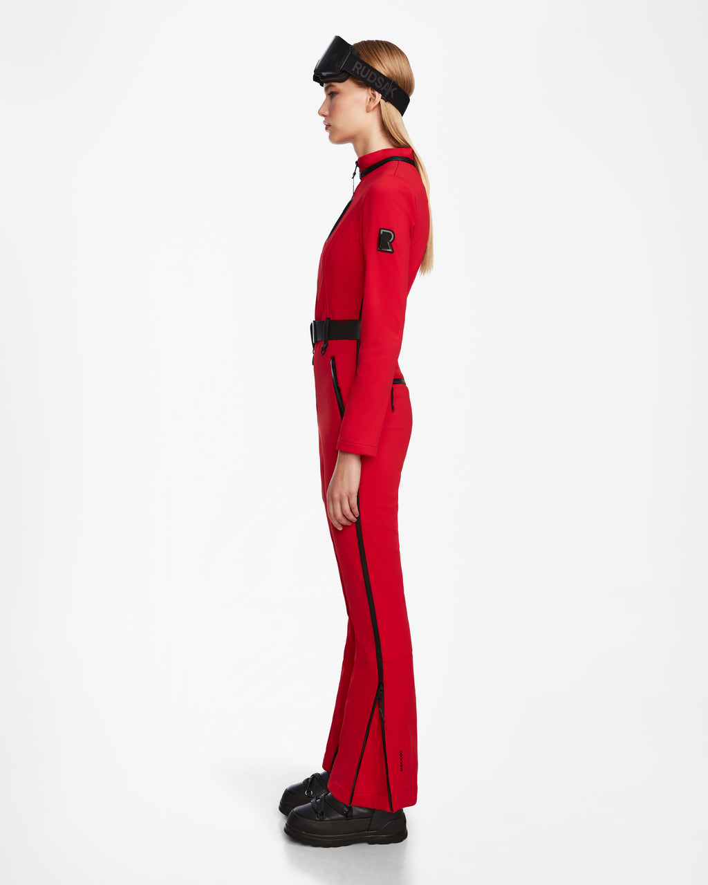 A woman stands in profile against a white background, wearing the Rudsak JOSIE - 8124817 Flame Red fitted ski suit with black zippers and belt, paired with black platform shoes and a black visor headband.