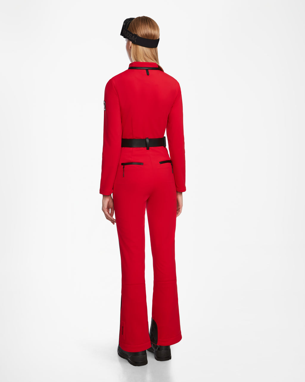 A person stands facing away, wearing the Rudsak JOSIE - 8124817 Flame Red fitted one-piece ski suit with black accents and zippers, black boots, and a black visor, set against a plain white background.