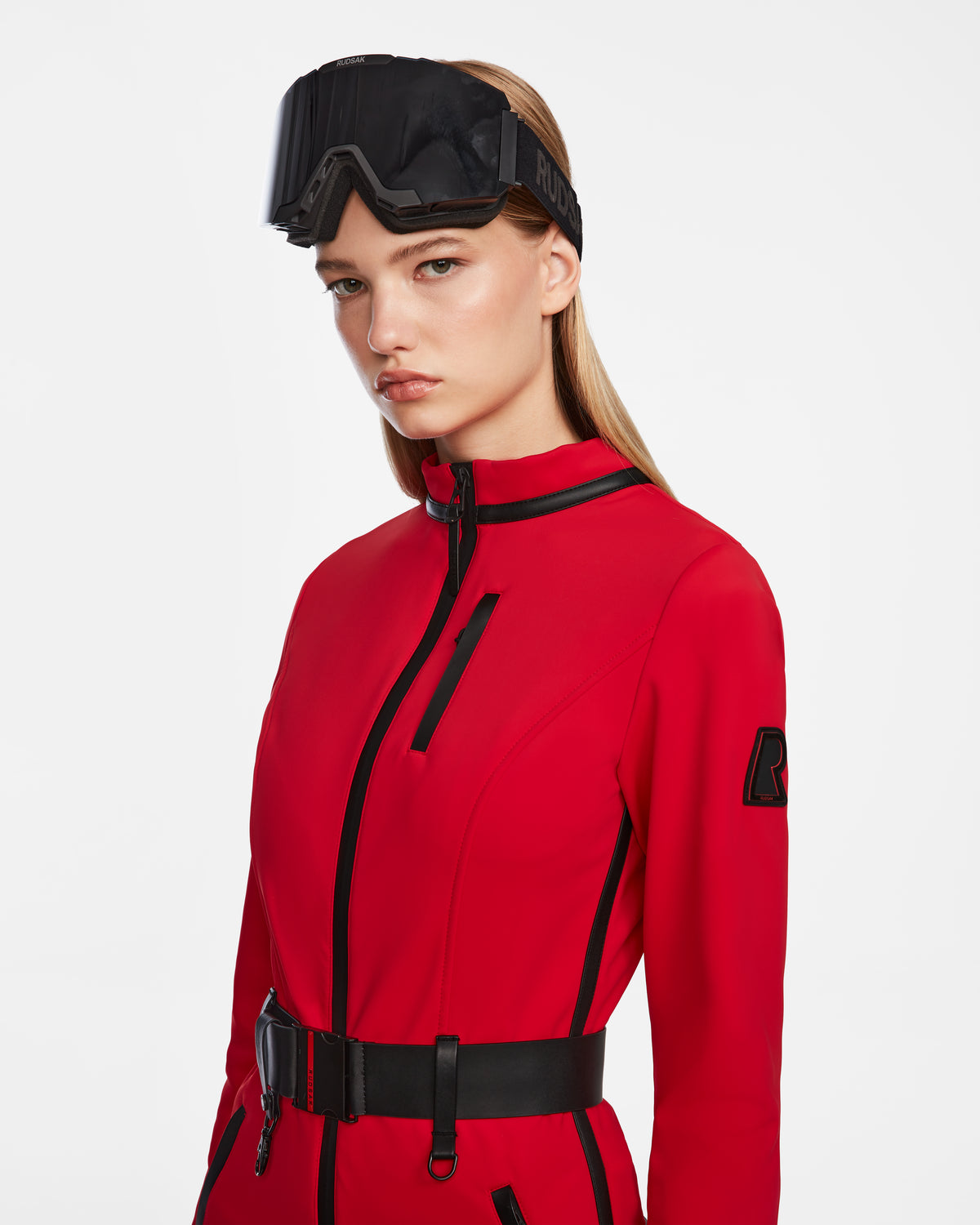 A woman wears the Rudsak JOSIE - 8124817 Flame Red one-piece ski suit with black accents and a black belt, featuring long straight hair and black ski goggles on her forehead, standing against a white background.